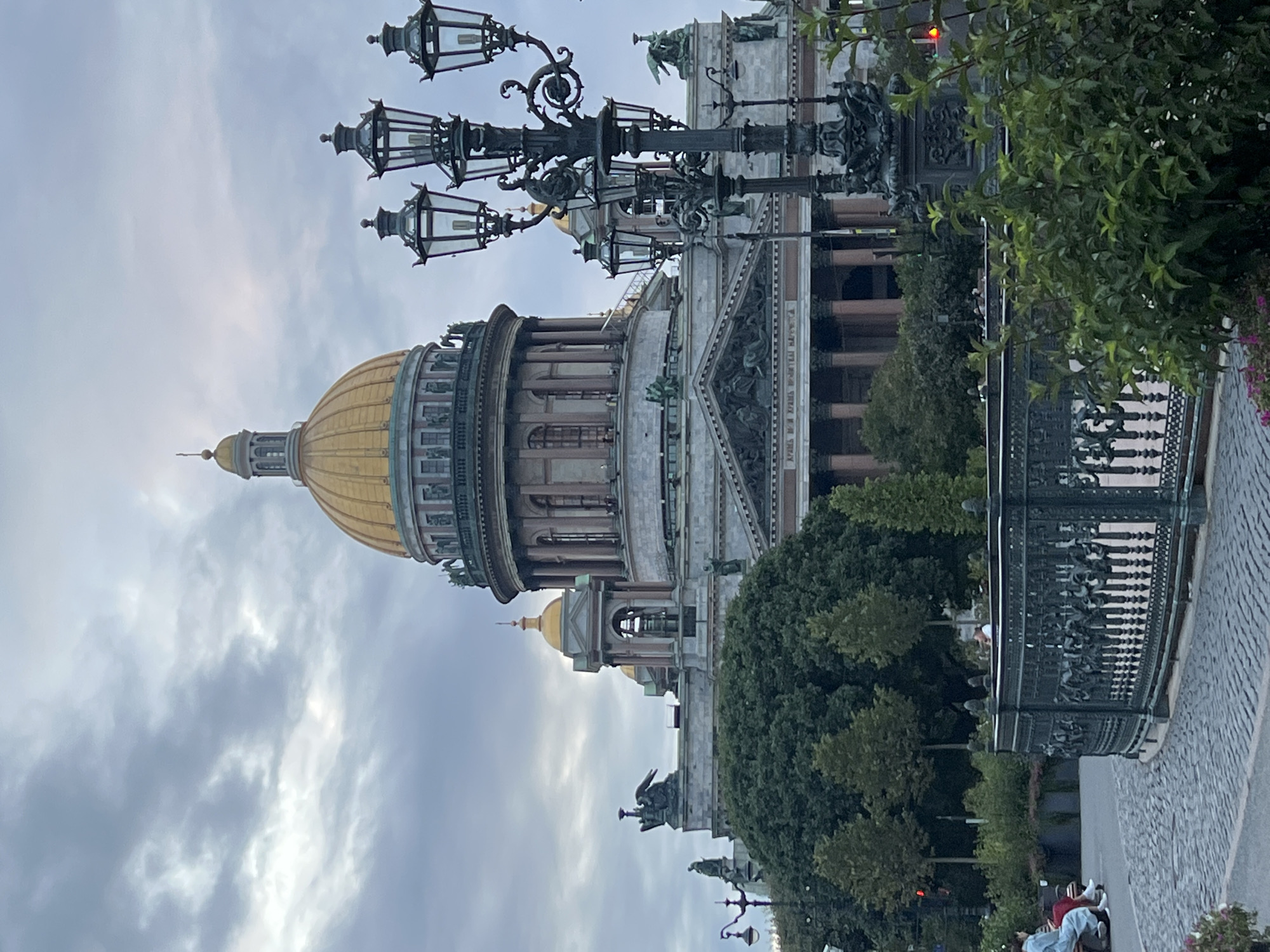 The image captures a view of St. Isaac's Cathedral in Saint Petersburg, Russia, under a cloudy sky. In the foreground, a decorative black metal fence encloses a public space with a cobblestone pathway. A multi-headed lamppost, characteristic of historic European street lighting, stands to the right of the cathedral, its ornate design contrasting with the imposing architecture behind it. Lush green trees and foliage surround the base of the cathedral, softening its grand stone facade. In the middle ground, a few individuals are visible, some seated on benches and others walking. The cathedral itself is a massive structure with a prominent golden dome, surrounded by smaller domes and architectural details, including statues and columns. The overall scene depicts a public square or park area adjacent to a significant historical landmark.