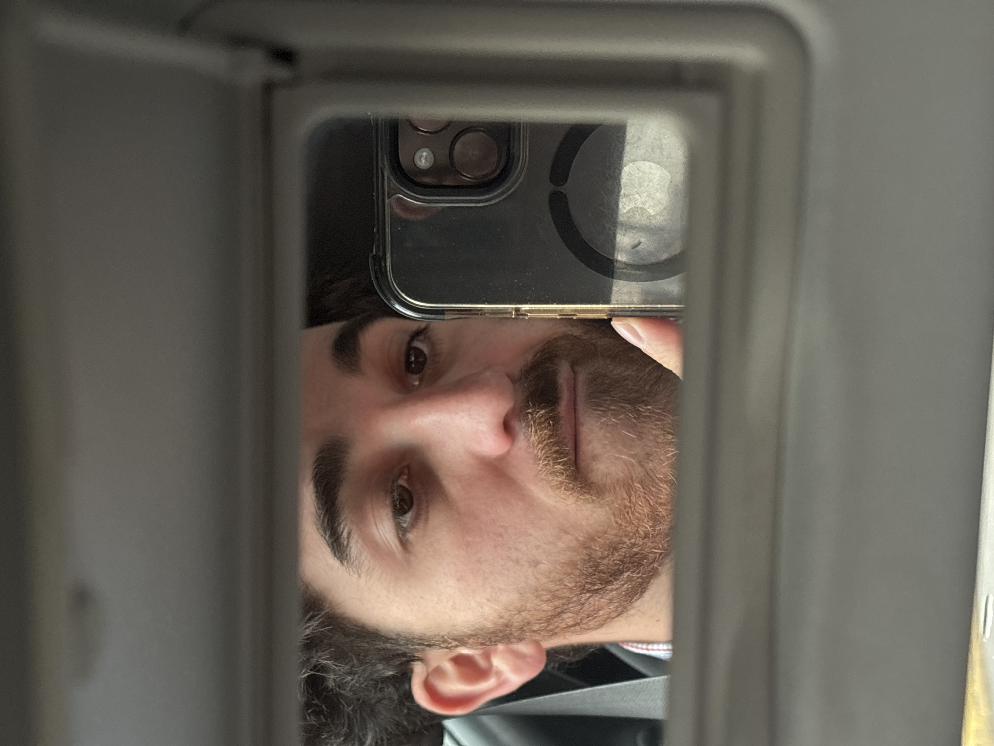 A man takes a selfie in a rearview mirror. The background is blurry and indistinct, making it impossible to determine the location as Puebla, Mexico. The man is holding a smartphone with a clear case, partially obscuring his face. He has dark hair, a beard, and brown eyes. He is wearing a black shirt.