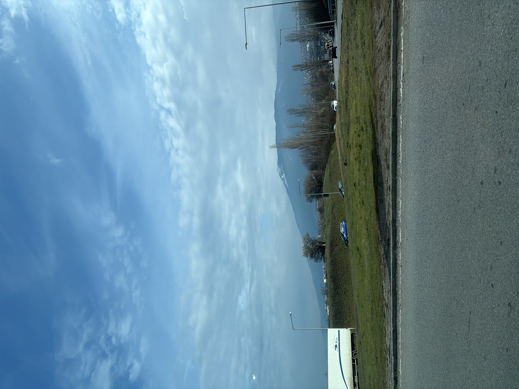 The image shows a view from a highway, possibly taken from a moving vehicle, with the landscape of Sofia, Bulgaria, visible in the background. To the left of the road is a green embankment with street lamps, and a blue car parked on the grass. In the distance, a mountain range is visible under a cloudy sky. Some buildings can be discerned along the base of the mountains. The highway pavement is visible on the right side of the image. A semi-truck partially visible in the image.