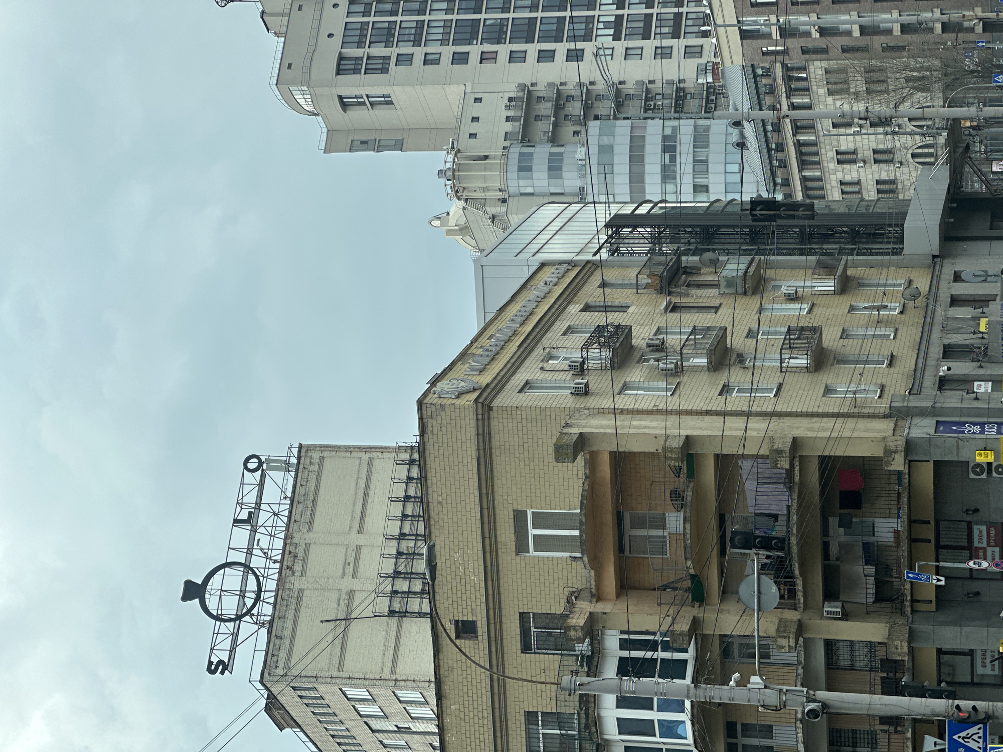 The image depicts a street scene in Kyiv, Ukraine. Several buildings of varying heights and architectural styles are visible. The building in the foreground has a distinctive rooftop sign featuring a stylized wedding ring with the word "SOLO" below it. The buildings are constructed with light-colored brick and concrete, and some have visible air conditioning units. A network of overhead power lines crisscrosses the scene. A traffic light pole and street signs are present at a crosswalk. Clothing is hanging from balconies. The sky is overcast.