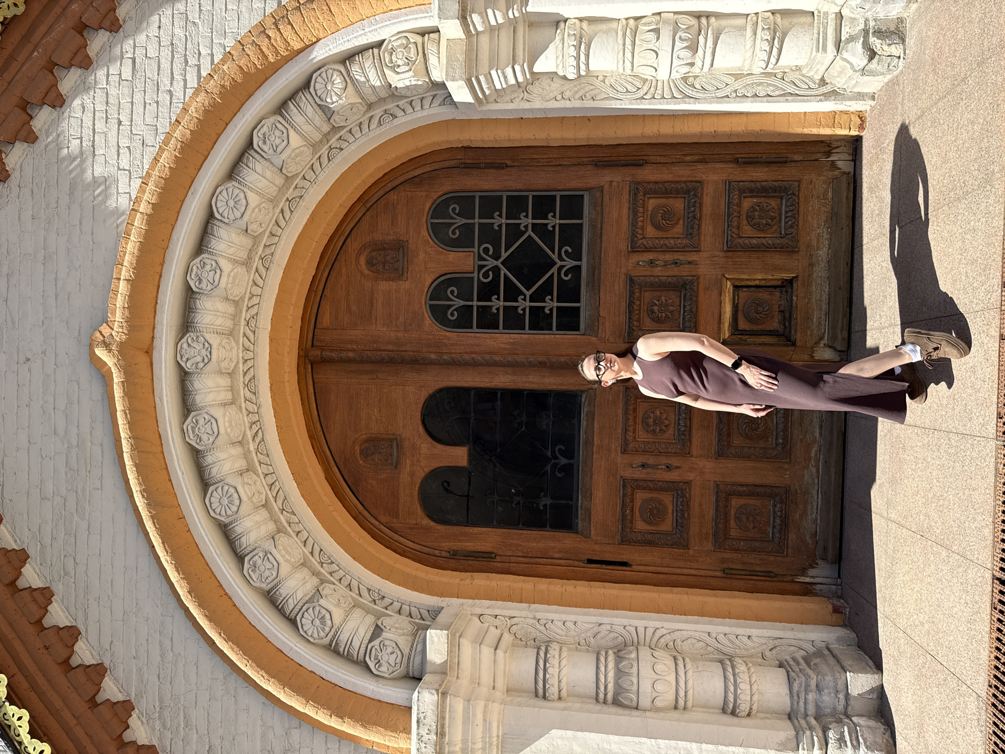 A vertically oriented medium shot depicts a woman in a long brown dress and brown shoes standing in front of a large, ornate wooden door. The door is set within an arched frame featuring decorative molding and is part of a light-colored building. A shadow of the woman is cast on the ground to her right. The image location is specified as Moscow, Russia.