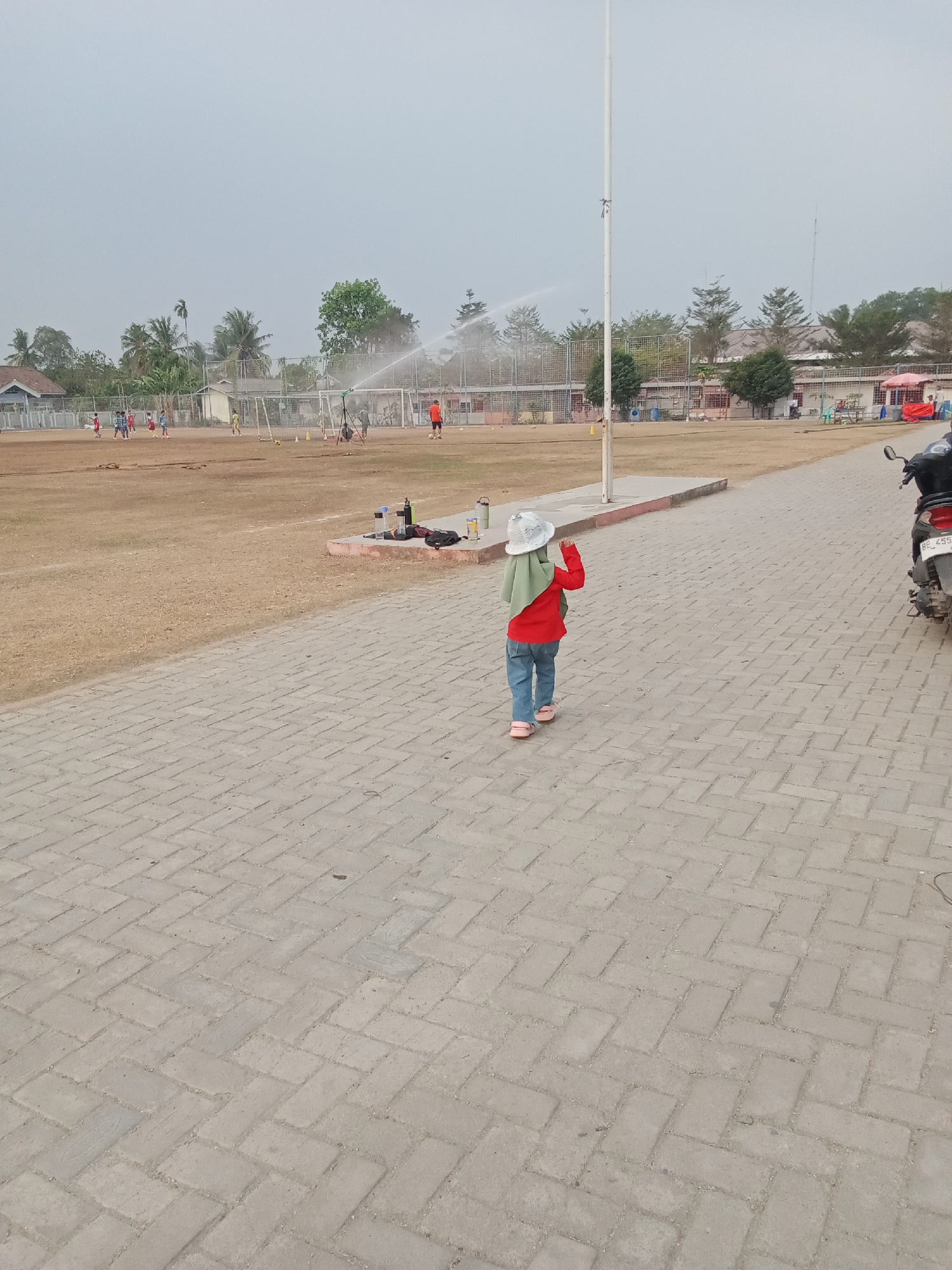 In Toto Katon, Indonesia, a child wearing a green hijab, a red long-sleeved shirt, blue jeans, and pink sandals is walking away from the camera on a paved walkway. The child is looking towards a soccer field where a sprinkler is watering the grass. Several children in soccer uniforms are visible on the field, along with a few goalposts. A white flagpole stands near the walkway. In the background, there are buildings and trees. A motorcycle is parked on the right side of the image.