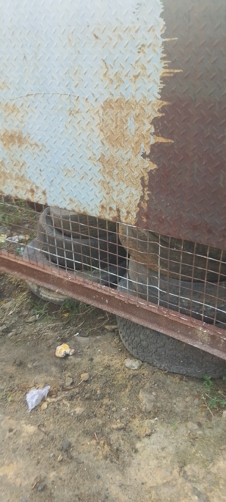 The scene shows a metal structure with a diamond plate pattern, heavily rusted, with a rusty metal cage at the bottom. Inside the cage, multiple tires are stacked. The ground is dirt with some debris, including a plastic bag and what appears to be food waste. The location is Unknown, Unknown.