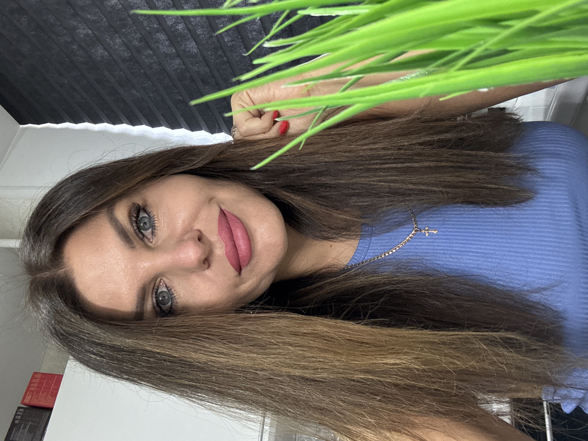The image shows a close-up, head-and-shoulders portrait of a woman with long, brown hair, blue eyes, and pink lipstick. She is wearing a blue ribbed top and a gold cross necklace. In the foreground, a cluster of bright green blades of grass is visible, obscuring part of her right side. Behind her, a dark, textured surface is visible on the right, and a white surface with some red and black rectangular objects is on the left. The scene appears to be indoors, with some lighting that creates a subtle shadow on the right side of her face. There is no discernible information to place this image in Chaikovsky, Russia.