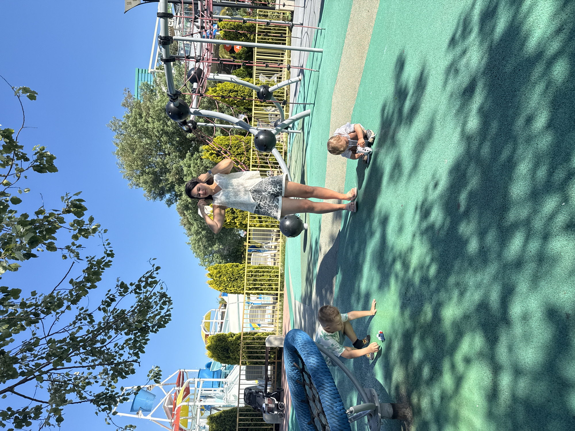 This image depicts a sunny day at an outdoor playground, possibly located in Sochi, Russia, given the presence of recreational facilities. In the foreground, two young boys are engaged in play on a green rubberized surface. One boy is crouched down, seemingly examining a small toy. The other boy is also kneeling, focused on a similar toy in front of him. Behind them, a woman stands with her hands near her head, appearing to be adjusting her hair or clothing. She is wearing a light-colored top and patterned shorts. The playground equipment is visible in the background, including climbing structures with ropes and poles, and in the far left background, what appears to be part of a water park with colorful slides and buckets. A metal fence separates the playground from a row of manicured evergreen bushes. The scene is characterized by bright sunlight casting dappled shadows from overhead trees onto the playground surface. A stroller is visible to the left, suggesting a family outing.