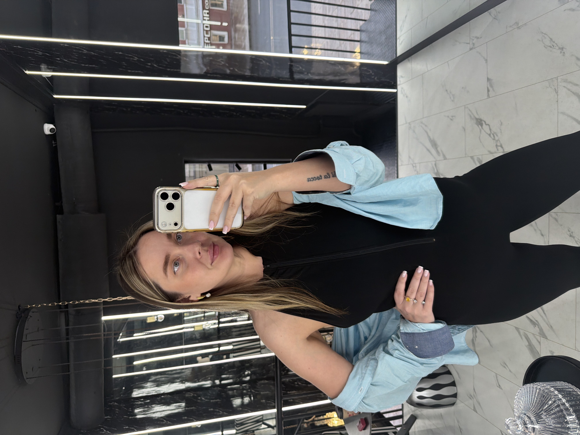 A woman with light-brown hair and blue eyes is taking a selfie in what appears to be a retail space. She is wearing a black top with a zipper and a light-blue shirt over it. She is holding a white and gold-trimmed cell phone in her left hand, which has a ring on the index finger. Her right hand, also adorned with rings, rests on her side.
The background reveals an interior with black walls illuminated by horizontal light strips. The marble flooring suggests a modern, upscale setting. A surveillance camera is visible in the upper-left corner. A building with the inscription "ЕРСОНА 600" in Cyrillic letters is visible through an upper window, likely placing this scene in Moscow, Russia.