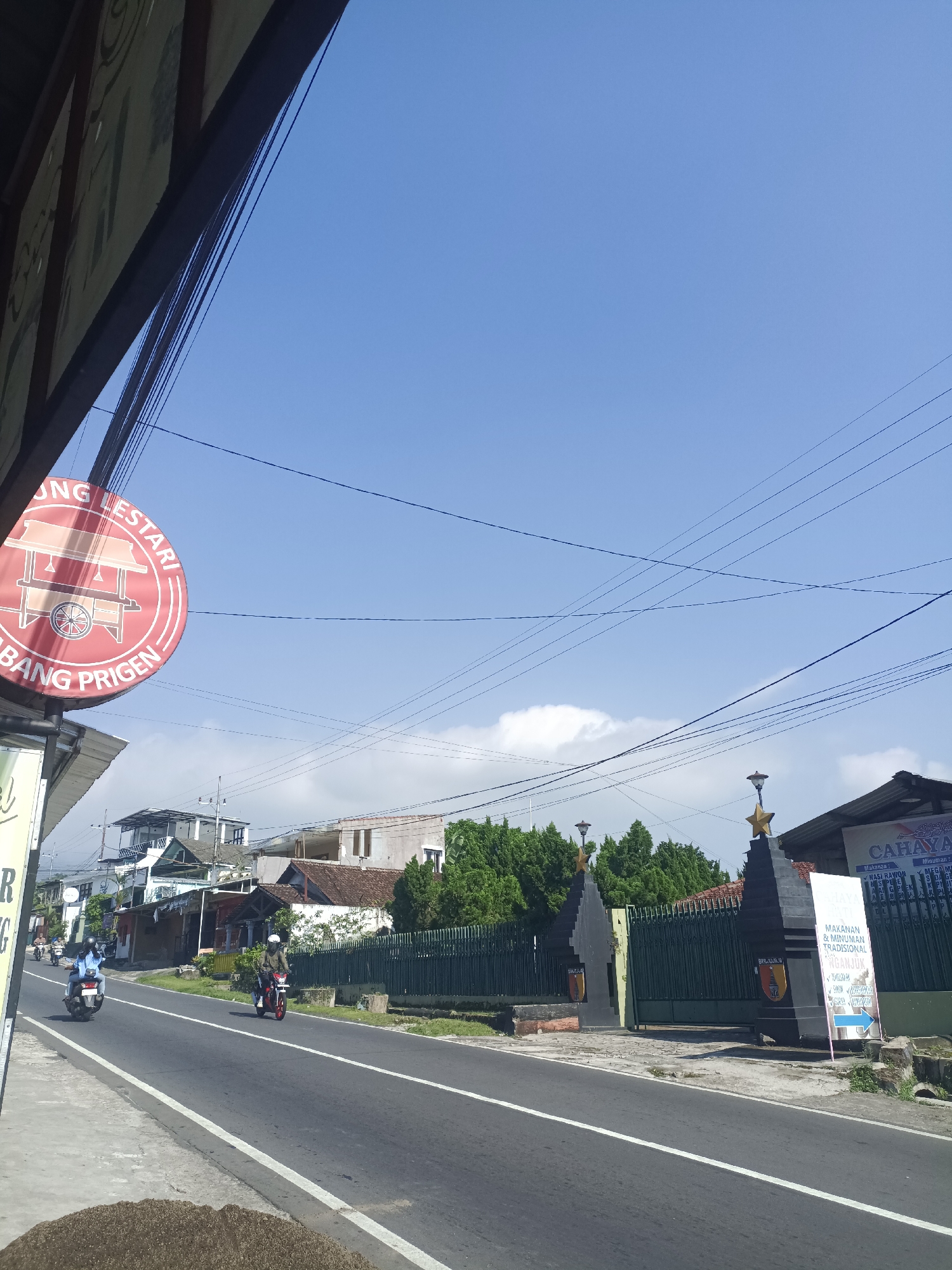 The scene depicts a street in Prigen, Indonesia, under a clear blue sky with power lines overhead. The road is asphalt, with a solid white line dividing the lanes. Two motorbikes are traveling on the road. The left side of the frame shows the corner of a building and a sign reading "UNG LESTARI ABANG PRIGEN." In the background, houses are visible, and the right side shows a building and a sign saying "CAHAYA MAKANAN & MINUMAN TRADISIONAL PRIGEN."