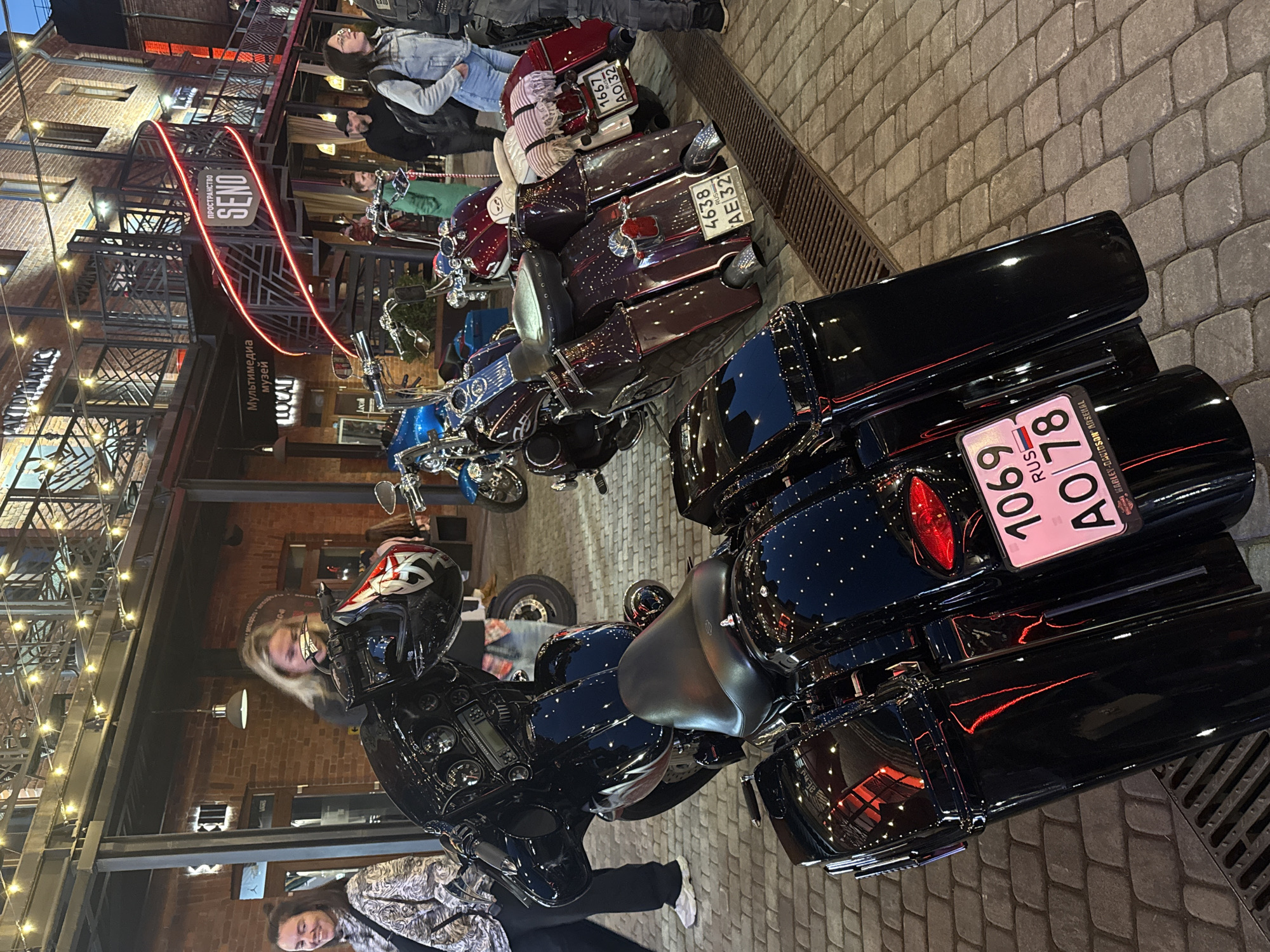 The image captures a nighttime scene on a cobblestone street in Saint Petersburg, Russia, likely in an entertainment or cultural district given the illuminated building facade. The street is lined with motorcycles, primarily Harley-Davidsons, parked in front of a multi-story building with a contemporary architectural style featuring exposed brick and metal accents. Neon signs with Russian text are visible, including "ПРОСТРАНСТВО SENO" (SENO SPACE) and "Мультимедиа музей" (Multimedia Museum). Several individuals are present, some walking and others near the motorcycles. One woman in the foreground is partially obscured by a black motorcycle, while another woman on the left is smiling. The motorcycles are detailed and appear to be in good condition, some adorned with luggage. The overall atmosphere suggests a gathering or event centered around motorcycles, possibly at a venue or exhibition.