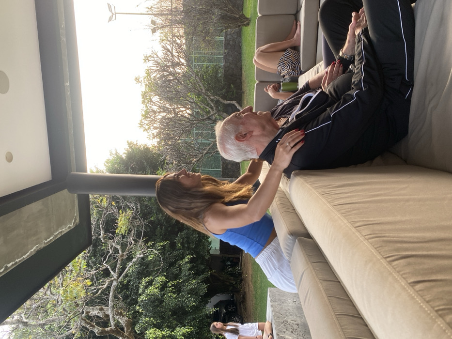 Outdoors, a woman in a blue top and white pants attends to a seated older man in a black jacket and pants with white stripes. The man rests on a light-colored outdoor sofa. Another person's legs are visible in the background, also seated on the sofa. Trees and foliage fill the background, with a glimpse of a white tennis court fence. An additional figure, smaller in scale, stands below the woman and the man.