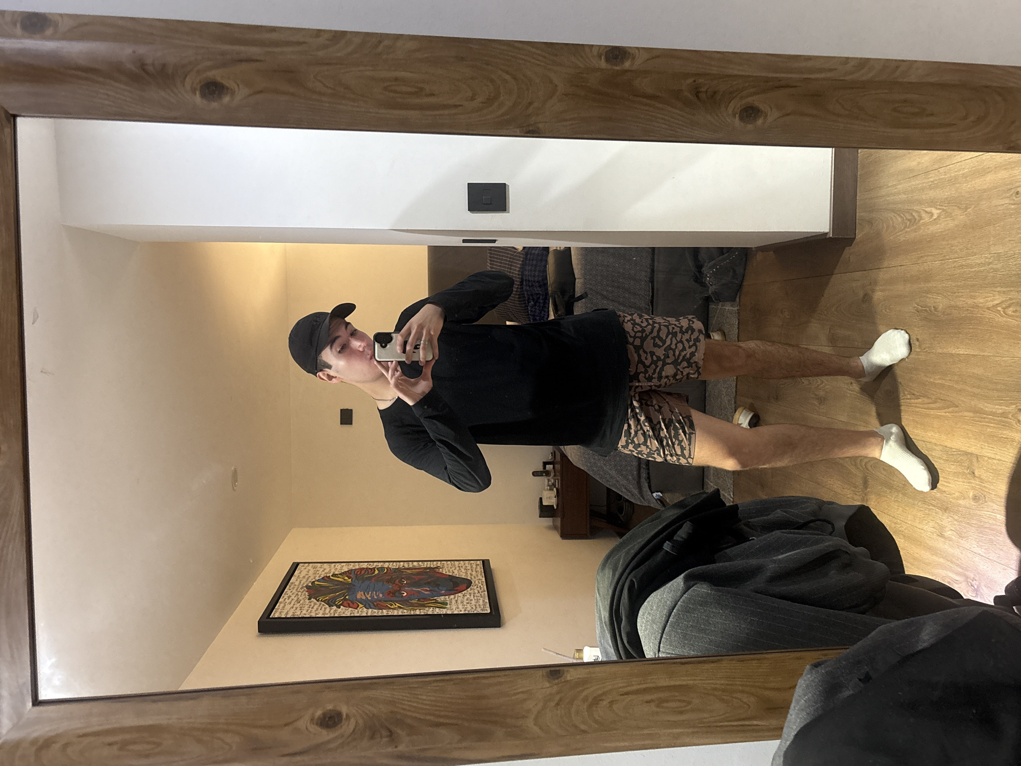 A full-length mirror shows a man taking a photo of himself with a cell phone. He is wearing a black long-sleeved shirt, patterned shorts, a black baseball cap, and white socks. The room behind him contains a painting on the wall, a dresser, and furniture. The floor appears to be wood laminate. The location of the city of San Bernardino Tlaxcalancingo, Mexico is not discernible from the image.