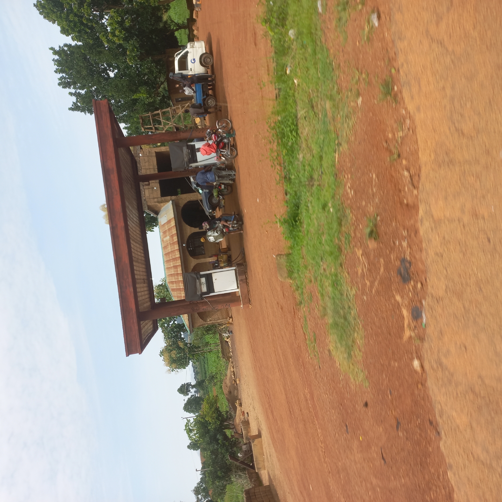 In Dorowa, Nigeria, a rural gas station is depicted under a partly cloudy sky. The station features a covered canopy with a corrugated metal roof, supported by red-brown pillars. A white fuel pump with a black screen stands under the canopy. Several people are present, including two individuals on a red motorcycle, one wearing a white helmet and a dark jacket with the number "09" on the back, and another in a red hooded sweatshirt. To the right, a white truck with a blue cargo bed is parked, with several people gathered around it. A ladder is leaning against the building behind the truck. The ground is covered in red-brown dirt and patches of green grass. In the background, trees and vegetation are visible, with some farm fields in the distance. The overall scene suggests a functional, albeit rustic, stop for fuel and transport in a rural Nigerian setting.