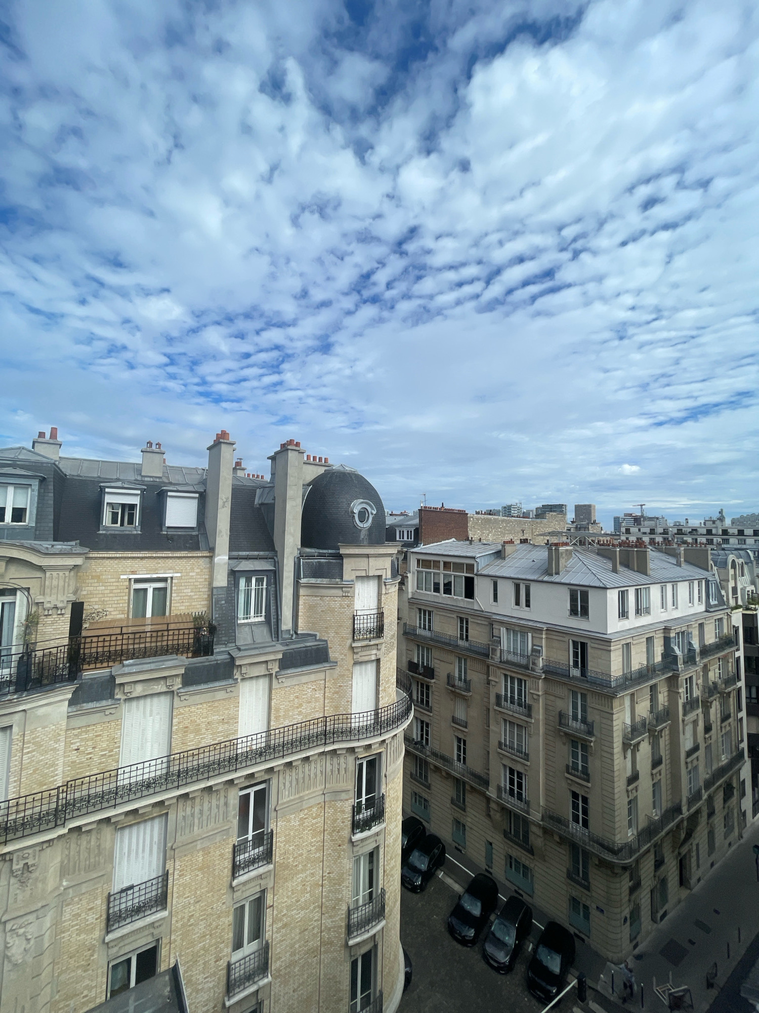 This is an aerial, eye-level, wide shot taken from an elevated position, looking down upon a Parisian cityscape. The foreground features the light-yellow brick facade of an elegant Haussmannian building with an arched, curving side. The building has multiple stories with balconies that feature black wrought-iron railings. The windows are numerous and many are covered by white rolling shutters. The roofline is complex, with various dormer windows and pitched slate roofs, punctuated by several red-capped chimneys. One prominent feature is a dark, domed turret on the corner of the building.
To the right of the foreground building, another similar Haussmannian building stands, this one with a more neutral, beige facade. It also has multiple stories, balconies with railings, and many windows. This building is rectangular and forms a street corner. Parked on the street below this building are several dark-colored cars, including a few black sedans and SUVs.
In the background, a dense collection of urban buildings stretches across the horizon. These buildings vary in height and architectural style, with some appearing more modern with flat roofs and larger windows. The sky above is a pale blue, filled with scattered, wispy white clouds that create a textured pattern. The overall impression is of a bright, overcast day in an urban environment. The scene is still and devoid of any discernible action or interaction between people.