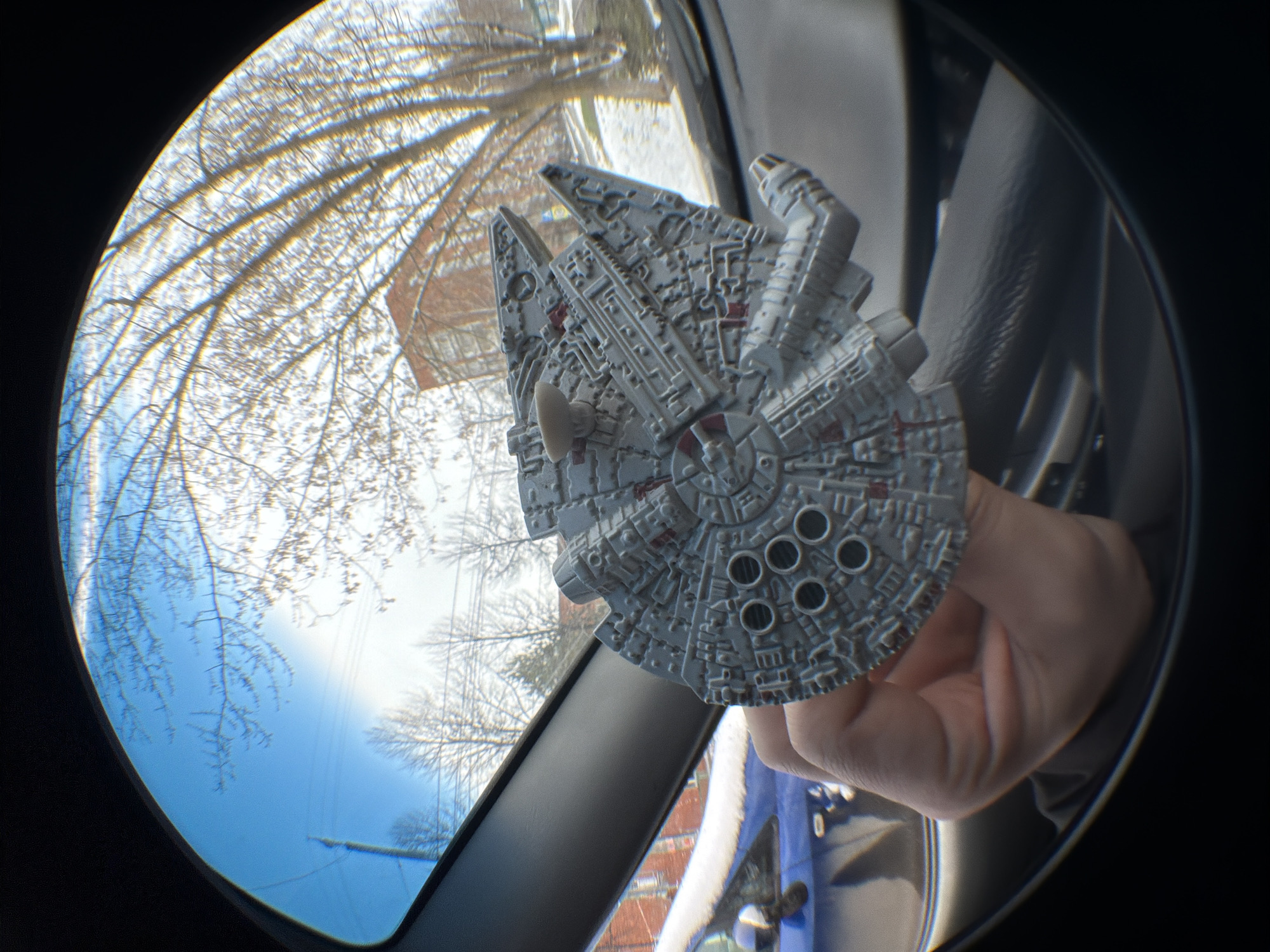 A fisheye lens captures an indoor scene where a gray Millennium Falcon toy is held by a hand. Outside the car window, bare trees suggest winter, with a view of a brick building and a blue sky. Due to the visual distortions from the lens, it is difficult to determine any particular location, and identifying the city as Izhevsk, Russia is not possible from the visible information.