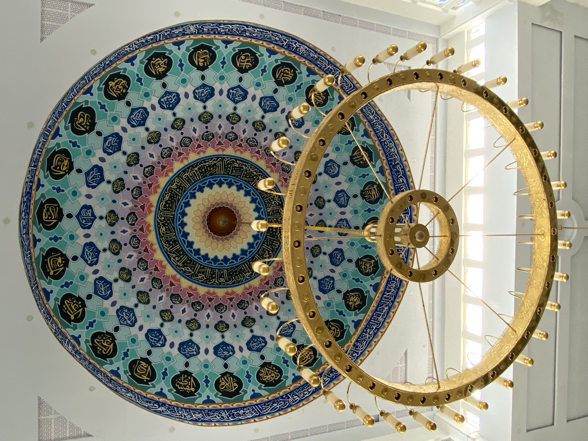 The media file captures an upward view of a mosque's interior in Lhokseumawe, Indonesia. The primary subjects are the ornate ceiling dome and a large, multi-tiered golden chandelier. The dome is decorated with intricate Islamic calligraphy and geometric patterns in vibrant colors, including blues, greens, purples, and yellows. The calligraphy appears to be Arabic script, common in mosque decorations.
The chandelier is circular and made of polished gold-colored metal, featuring numerous light bulbs or sockets with white casings. It hangs prominently in the foreground, partially obscuring the dome. The overall lighting suggests it is daytime, with natural light filtering in from large windows visible in the background, which are framed by white architectural elements. The windows are rectangular and segmented, indicating a modern or semi-modern architectural style for the mosque.
The scene is one of architectural beauty and religious devotion, conveying a sense of grandeur and peace. There is no visible activity or event taking place, as the image focuses on the static elements of the mosque's interior. The location cues of Lhokseumawe, Indonesia, place this within a Southeast Asian Islamic context. The image evokes a feeling of awe and reverence. No specific time of day is definitively indicated beyond the presence of daylight, and no weather conditions are discernible. The focus is purely on the static visual elements of the mosque's interior design.