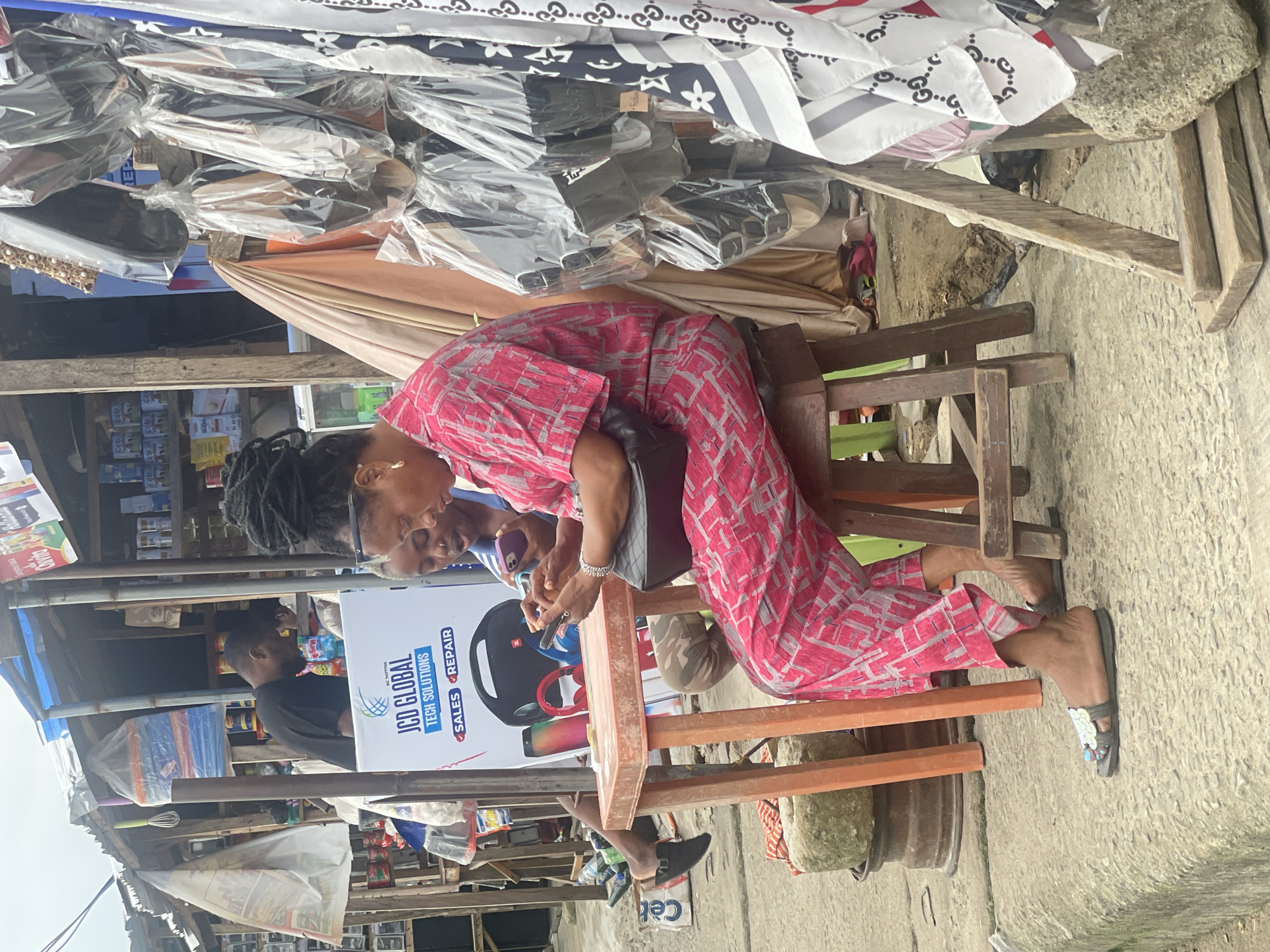 The image depicts an outdoor market scene in Ado, Nigeria, with a focus on three individuals. In the foreground, a woman wearing a pink and gray patterned outfit is seated on a wooden chair. She is looking down at her phone, which she is holding in her hands. To her left, another person, partially obscured, also appears to be looking at a phone. Behind them, a man in a black t-shirt is visible. The market appears to be stocked with various goods, including fabric, electronics, and household items, some of which are displayed on shelves and under tarps. The overall impression is a bustling marketplace.
A prominent sign on a white background displays the text "JCD GLOBAL TECH SOLUTIONS" with smaller text below indicating "SALES REPAIR". The image suggests it is daytime, with the lighting indicating it's not early morning or late evening. The concrete ground and the types of stalls suggest an urban or semi-urban environment. The expressions of the individuals are neutral, focused on their activities.
