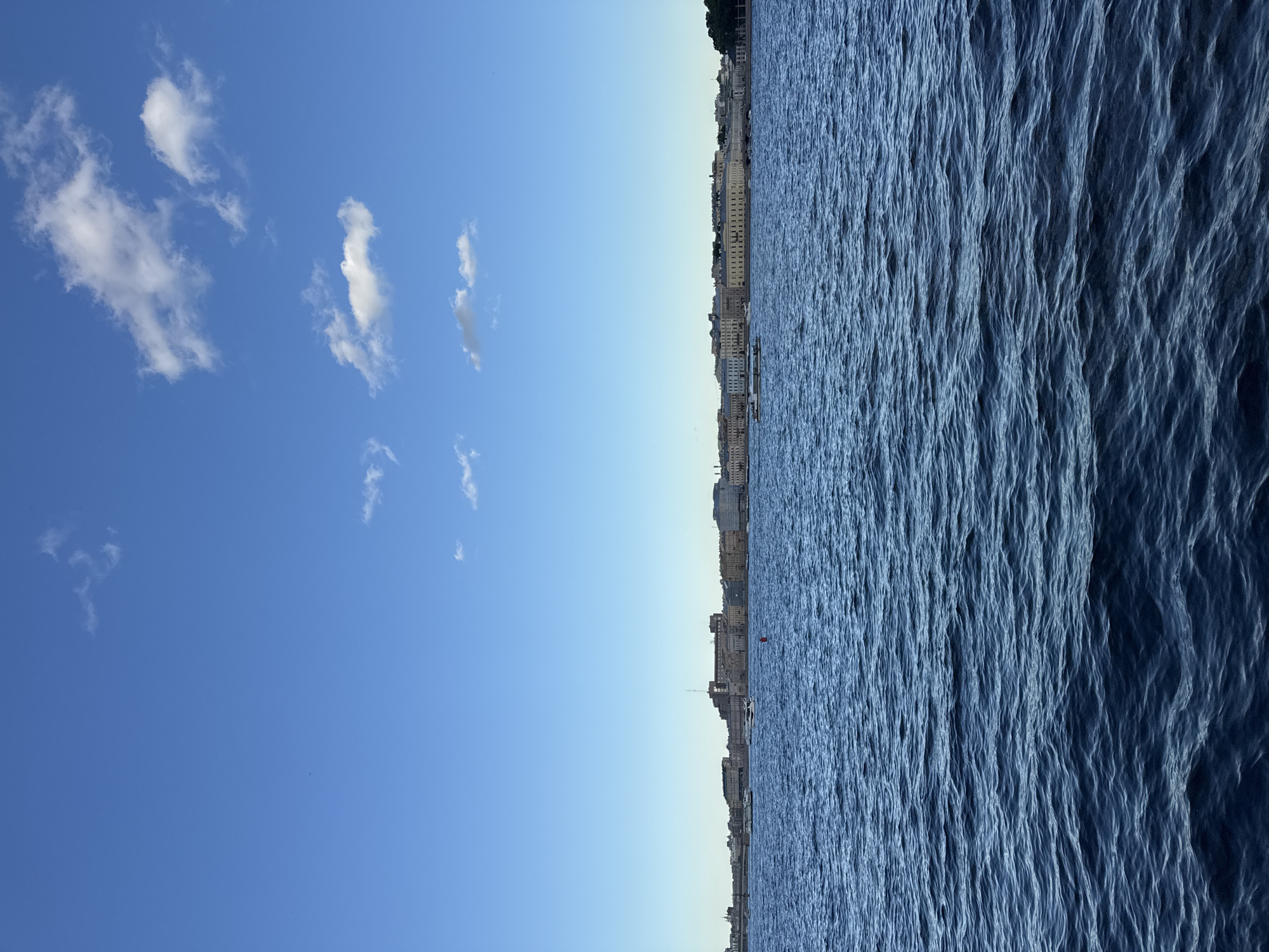 This is a photograph of the Neva River in Saint Petersburg, Russia, viewed from the water. The image is divided into two horizontal sections: the lower two-thirds depict the water, and the upper third shows the sky.
The Neva River is a deep blue and exhibits gentle waves, suggesting a slightly breezy day. Several boats are visible on the water, including what appear to be smaller pleasure craft and a larger vessel in the distance. A red buoy is also present in the middle ground.
Across the river, a long, continuous cityscape lines the horizon. The architecture is characterized by buildings with light-colored facades, many featuring classical design elements such as columns and ornate windows. The buildings are of varying heights, forming a consistent urban skyline. Hints of greenery, possibly trees, are visible at the far right edge of the scene.
The sky is a clear, vibrant blue with a few scattered white clouds. The overall impression is of a calm, sunny day in a historically significant city.