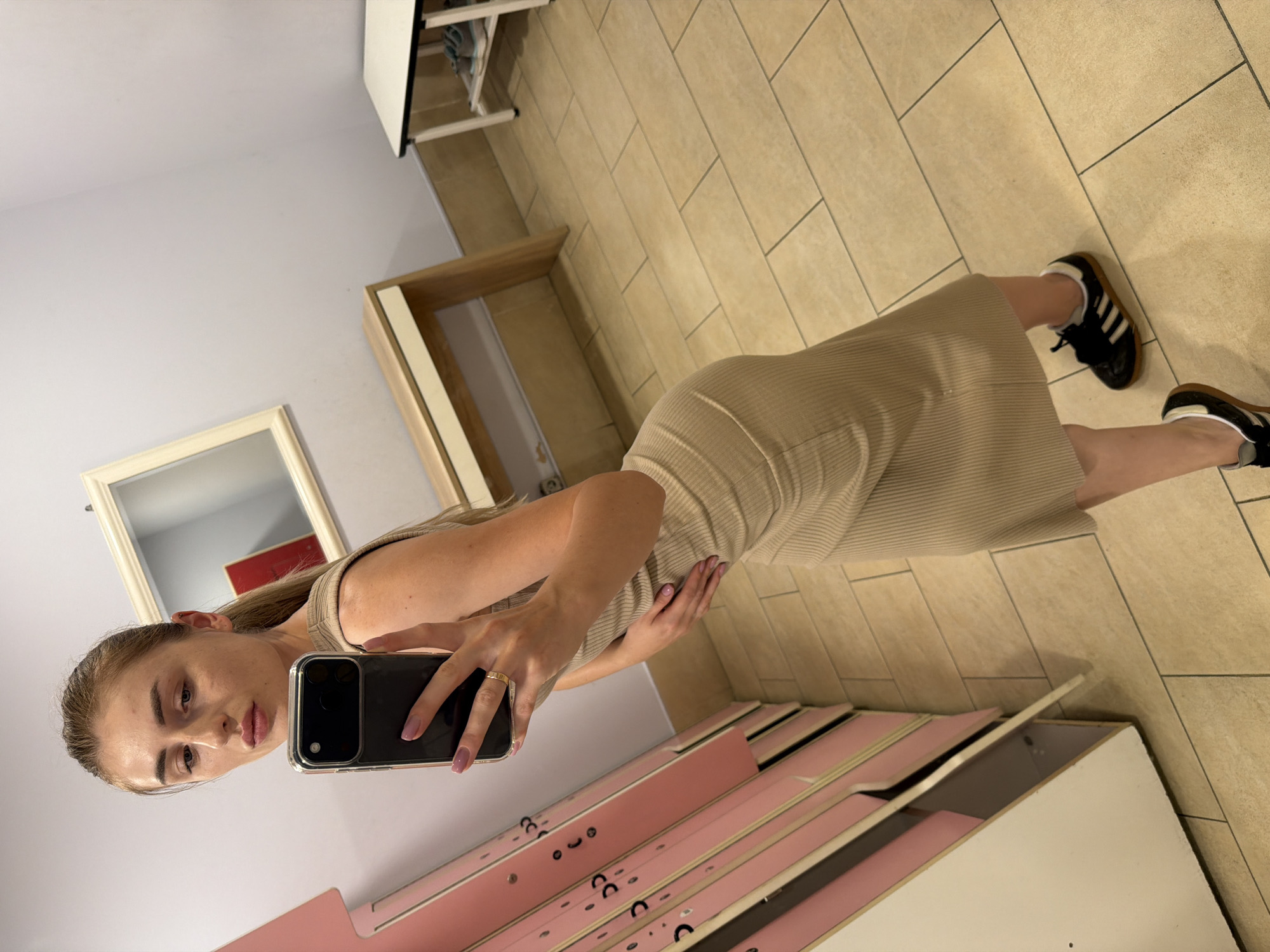A young woman with blonde hair pulled back is taking a mirror selfie in a room with light-colored walls and beige tiled flooring. She is wearing a beige ribbed knit dress and black Adidas sneakers with white stripes. The room appears to be a changing room or a storage area, with shelves and furniture visible in the background. In the foreground, she is holding a dark smartphone to take the picture. Her left hand rests on her hip, and her right hand is holding the phone. A gold ring is visible on her left ring finger. The lighting suggests it could be daytime. There is no visible text in the image.