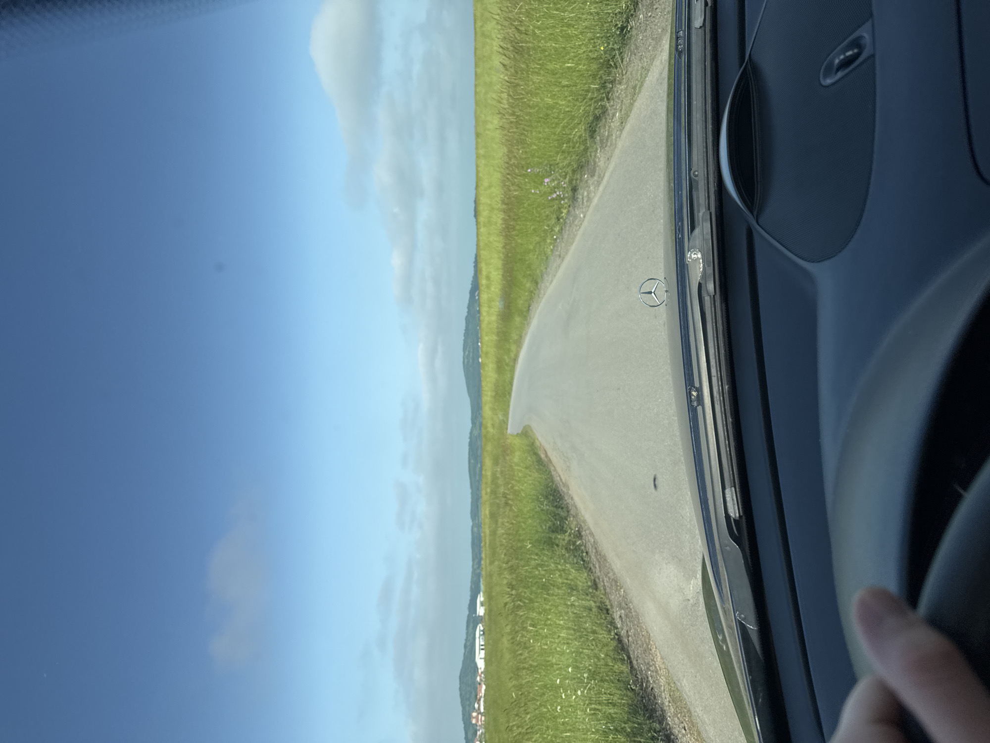 The image presents a first-person view from the interior of a Mercedes-Benz vehicle, situated in Veselie, Bulgaria. A light-skinned hand is visible grasping a dark steering wheel in the lower left corner, indicating the presence of a driver. The dark dashboard and the base of the windshield occupy the foreground, with a prominent silver Mercedes-Benz three-pointed star emblem affixed to the center of the hood. A textured speaker grille or compartment is visible on the passenger side of the dashboard. The vehicle is on a narrow, asphalt road that gently curves to the left as it recedes into the distance. The environment consists of expansive green fields of tall grass and wildflowers flanking both sides of the road. In the far distance, low-lying hills are visible, and to the left, a cluster of red-roofed buildings and several white structures are discernible. The sky above is a clear blue, adorned with scattered white cumulus clouds. Minor circular dust or debris spots are visible on the camera lens, primarily in the upper sky portion of the image.