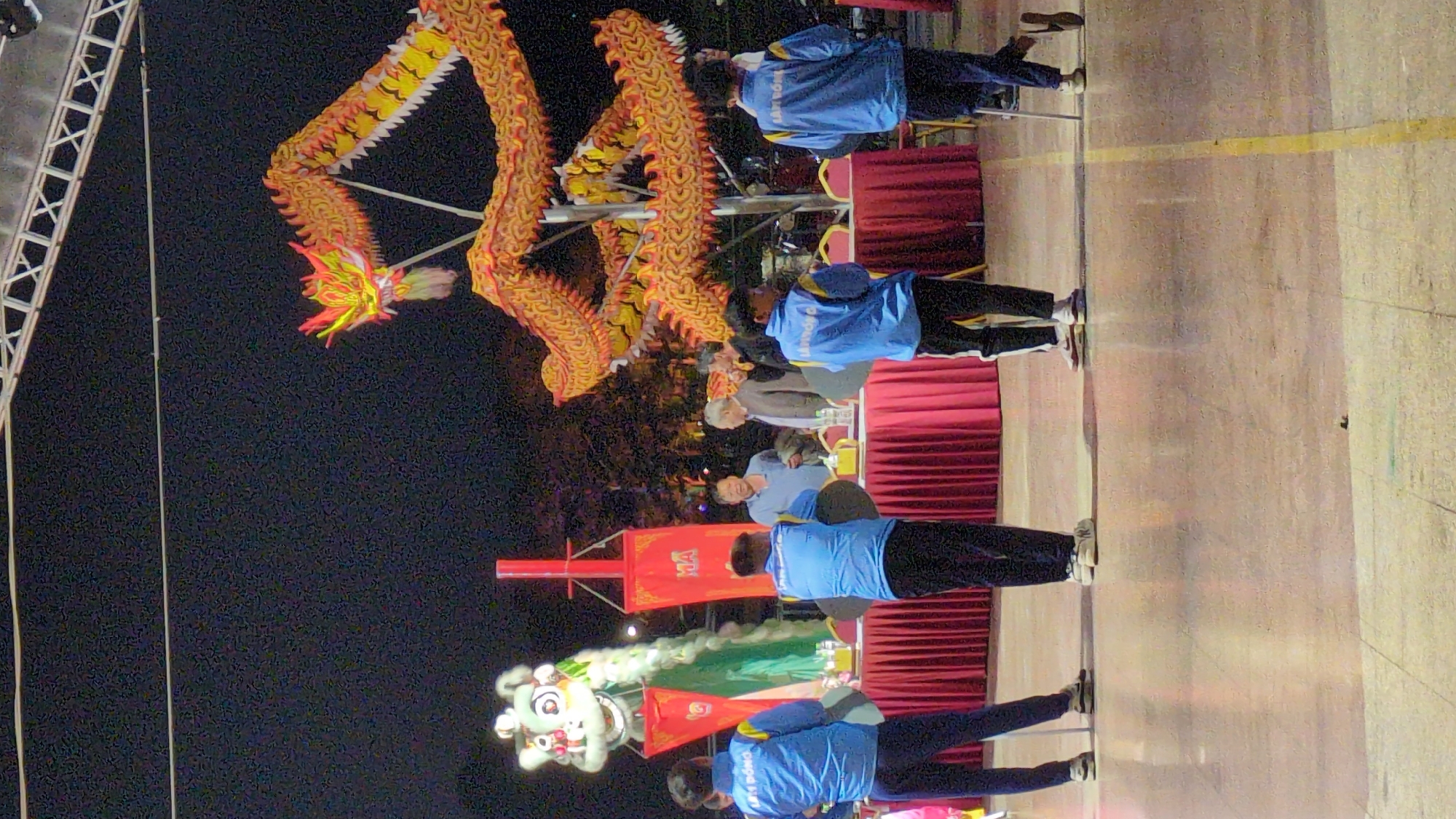 A horizontally-oriented photograph depicts a lion and dragon dance performance in Da Lat, Vietnam. The upper portion of the frame, set against a dark, possibly nighttime sky, features a dragon, its body rendered in shades of orange, yellow, and white. Positioned below the dragon is a lion costume.
A group of individuals, dressed in matching blue jackets, stand along the lower half of the image. The performers are shown in various positions: some standing, others in motion, suggesting they are participating in the dance. Additionally, individuals are seated at tables draped with red tablecloths.
The foreground is a light-colored surface. The image has been rotated 90 degrees.