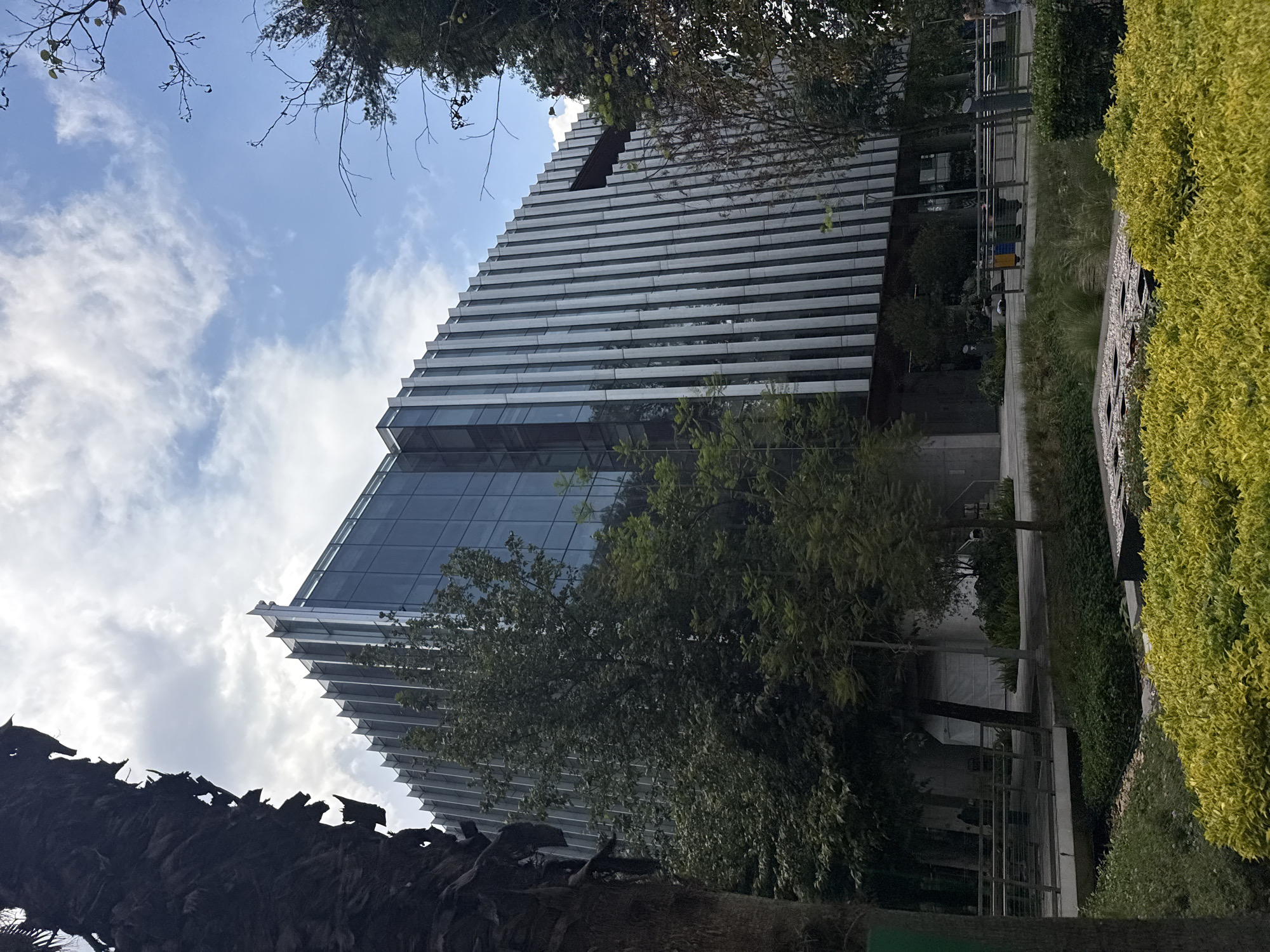 The image shows a modern building with a unique, staggered or stepped facade, possibly located in Mexico City, Mexico. The building has a combination of glass and white horizontal elements. The lower part of the building is predominantly glass, while the upper part has a pattern of alternating glass and white tiers. The sky above is partly cloudy. A variety of trees and foliage surround the building, with some branches obscuring the view of the building and the sky. A sidewalk or path is visible in front of the building, with some infrastructure like railings and benches. A dense hedge lines the path. The overall impression is that of a modern architectural structure integrated with green space in an urban environment.