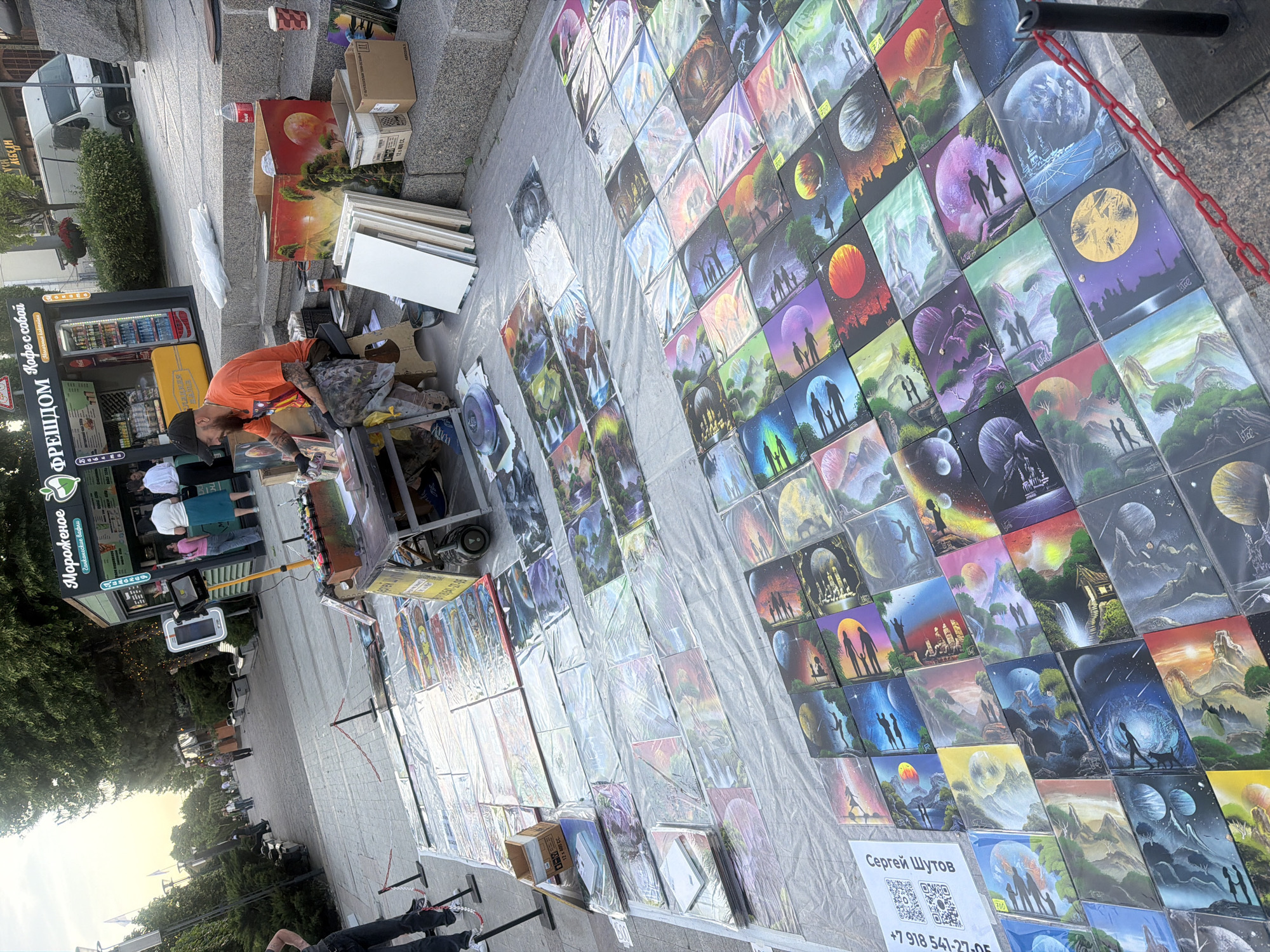 Here is a detailed description of the image:
**General Description:**
The image depicts an outdoor art market or street fair scene. A vendor is showcasing and selling artwork, primarily paintings, laid out on a large, white plastic sheet covering the ground. The scene is likely in a public urban area.
**Location Details:**
Based on the signage in the image, the location is likely in Rostov-on-Don, Russia. Signage written in Russian with "ФРЕШДОМ Кофе с собой" (FRESHDOM Coffee to go) and "Мороженое" (Ice Cream) can be clearly identified.
**People:**
1. **Vendor:** An adult male wearing an orange shirt and a cap is positioned near a cart/table. He appears to be organizing or displaying the artwork. He has tattoos on his arms.
2. **Customers/Passersby:** There are several indistinct figures in the background, suggesting people walking by or browsing the market.
3. **Ice cream store staff:** There are people inside the ice cream store.
**Subjects/Objects:**
1. **Paintings:** Numerous paintings are displayed on the white plastic sheet. These appear to be fantasy landscapes with recurring motifs like planets, silhouettes of people, trees, and waterfalls.
2. **Display Sheet:** The white plastic sheet covers the ground, providing a clean surface to display the artwork.
3. **Cart/Table:** A cart or small table is next to the vendor, possibly used for supplies or as a workspace.
4. **Art Display:** Some paintings are stacked against boxes and easels on the left-hand side.
5. **Coffee and ice cream kiosk:** The shop features signs written in Russian: "ФРЕШДОМ Кофе с собой" (FRESHDOM Coffee to go) and "Мороженое" (Ice Cream).
6. **Signage:** A sign with the name "Сергей Шутов" (Sergei Shutov) and a QR code, along with a phone number, indicates the artist's name and contact information.
7. **Chains:** Metal chains can be seen dividing the displays from the street.
8. **Boxes and Packing Materials:** Cardboard boxes and other packing materials are visible near the vendor, likely used for storing and transporting artwork.
**Actions/Interactions:**
* The vendor is actively engaged in managing the display, potentially interacting with customers.
* Passersby are walking by, some likely browsing the artwork.
**Environment:**
* The environment is a paved urban area, likely a pedestrian walkway or street.
* Greenery is visible, with trees and shrubs lining the walkway.
* A vending machine can be seen to the left of the vendor.
* The overall atmosphere is likely that of a bustling, outdoor market or fair.