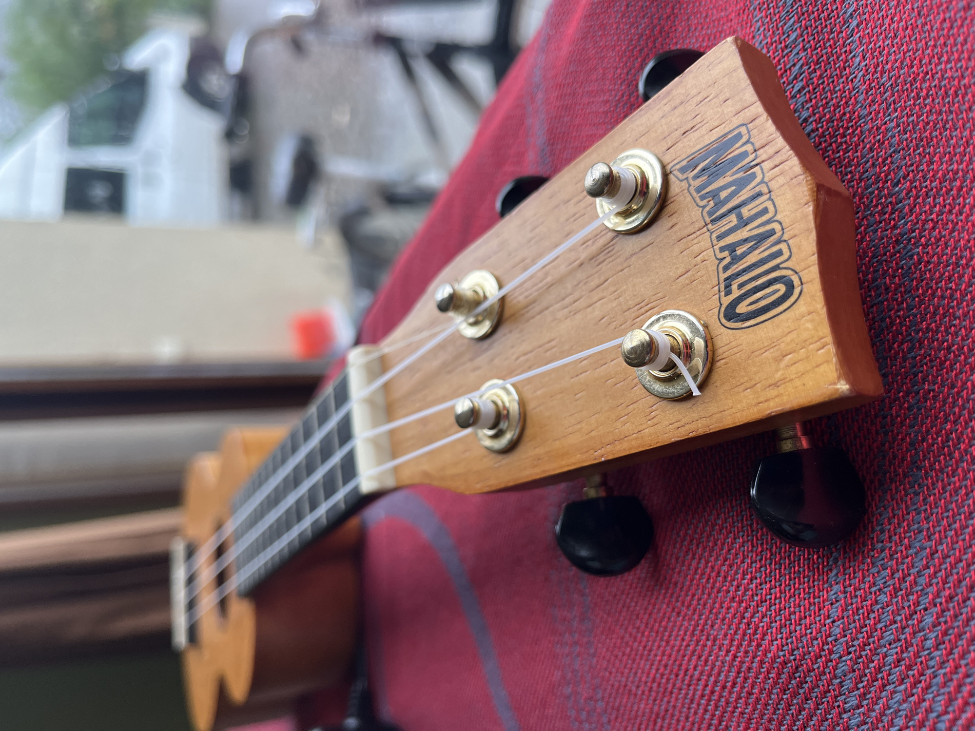 The image displays a close-up view of a ukulele's headstock, resting on a red and gray checkered fabric. The ukulele is made of light-colored wood and features four tuning pegs with black knobs. The brand name "MAHALO" is visible in black lettering on the headstock. The background is blurred, showing what appears to be a window or doorway with vertical blinds, and further back, a white van is partially visible, suggesting an outdoor or semi-outdoor setting. The scene is likely located in Ureki, Georgia, given the context of the request, though the image itself does not contain explicit location markers. There are no people or animals present in the frame.