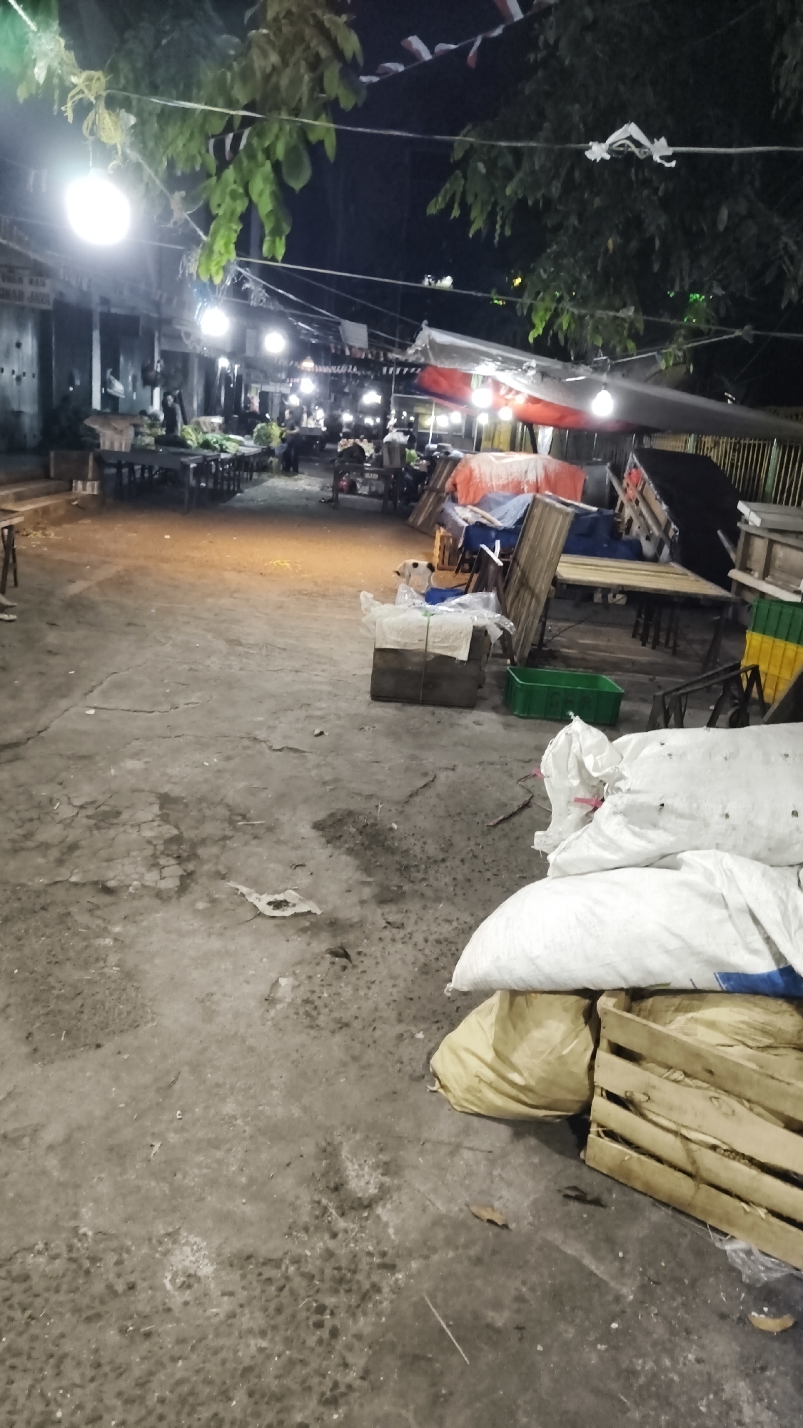 The image depicts a dimly lit outdoor scene, likely a marketplace or a food stall area, at night in South Jakarta, Indonesia. The ground is made of concrete with visible cracks and patches. In the foreground, several large white sacks and a yellow sack are piled next to a wooden crate. A green plastic container is also present. Further back, various market stalls with tables and chairs are set up, some covered by tarps and adorned with red and white flags, suggestive of a national holiday or celebration. Several people are visible, some seated at tables, while others appear to be vendors. The overall impression is one of a busy but somewhat unkempt nocturnal marketplace.