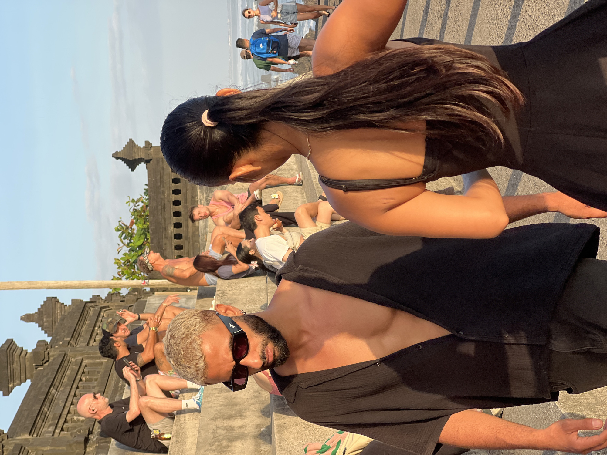 In North Kuta, Indonesia, a group of people are gathered on stone steps near the beach during golden hour. In the foreground, a man with blond hair and a beard, wearing a black open-neck shirt and sunglasses, is looking down. To his right, a woman with her back to the camera, her long dark hair tied in a ponytail, wears a black dress. Behind them, several individuals are seated on the steps, some holding drinks and phones. Further back, people are walking on a path towards the ocean, with waves visible in the distance under a clear sky. The architecture suggests a temple or a similar traditional structure.