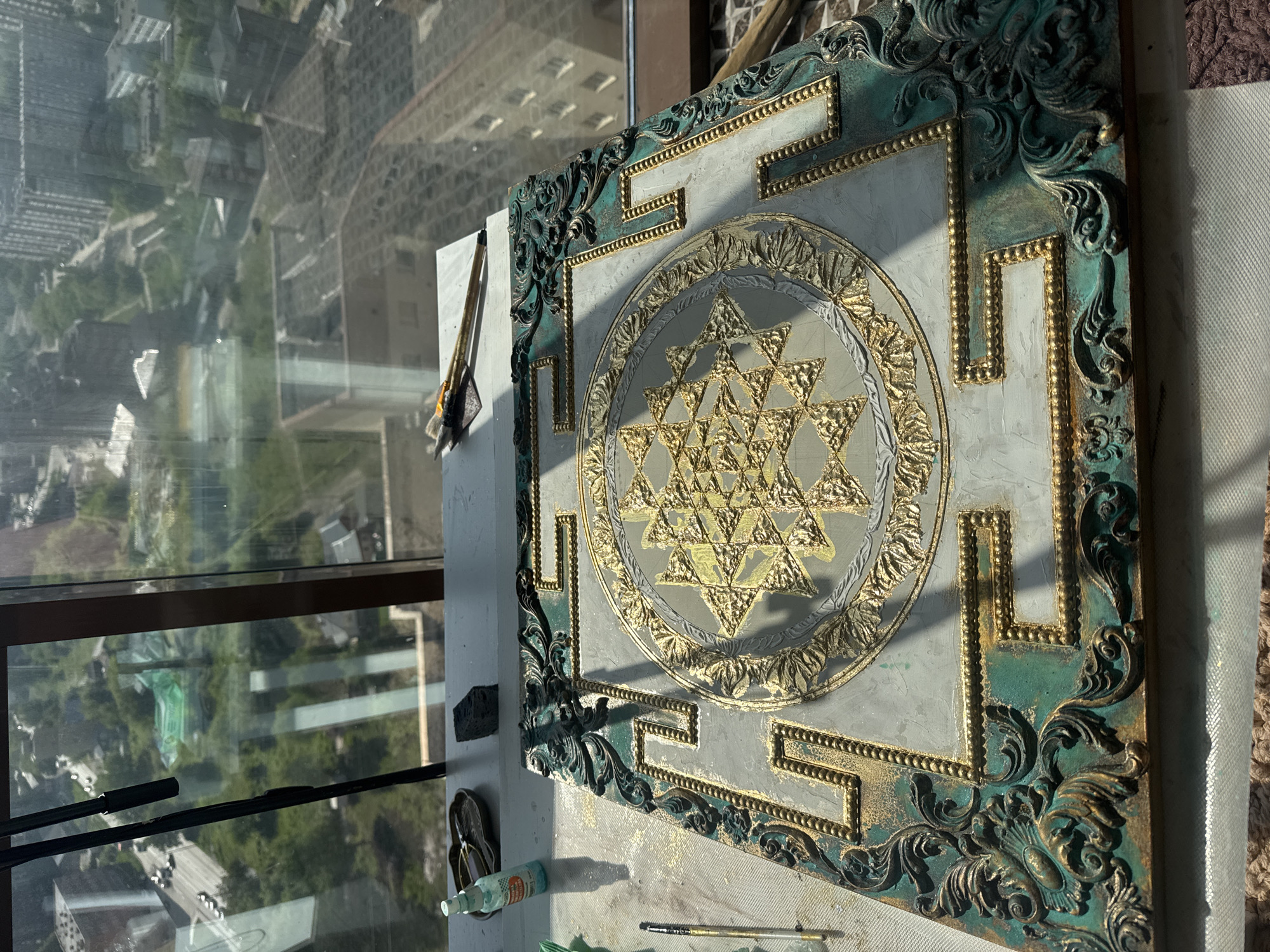 A highly ornate artwork, possibly a relief or painting of a Sri Yantra, is the central subject, illuminated by bright sunlight. The piece features a complex geometric pattern rendered in shimmering gold and silver-grey, set against a textured light grey background. It is framed by a rich, distressed teal-green border with intricate Baroque-style gold and dark green flourishes, creating an aged, luxurious aesthetic. Strong shadows are cast across the artwork and the surface it rests on, indicating a daytime setting.
The artwork is positioned near a large window, offering a high-rise perspective of an urban landscape, likely in Novosibirsk, Russia. Through the glass, multi-story buildings, green spaces, and roads are visible, partially obscured by reflections on the windowpane.
On the surface beside the artwork, several artist's tools are present, including multiple paintbrushes (one bundle secured with yellow tape), suggesting an ongoing or recently completed creative endeavor. A small, light blue bottle, a dark sponge-like object, and part of a flip-flop are also visible, indicating a casual workspace. No discernible text appears in the image.