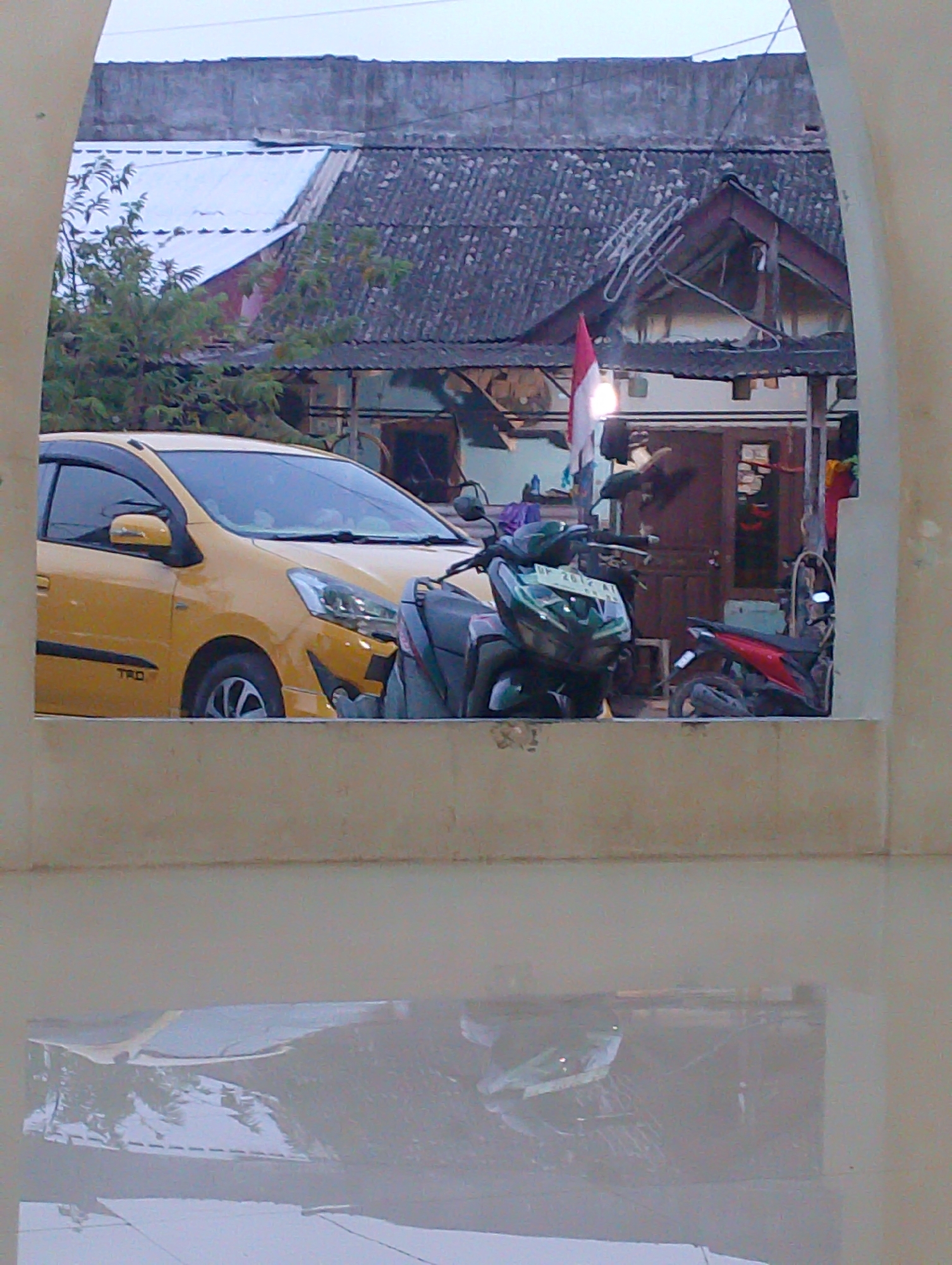The image, taken in Batam, Indonesia, depicts a street scene viewed through an archway. In the foreground, a pale beige wall runs horizontally, with a reflection of an outdoor scene visible on its surface. Beyond the wall, a yellow car is parked next to two motorcycles. The car has its driver's side facing the camera, and appears to have the "TRD" logo on its door. The motorcycles are positioned in front of a building with a dark brown door and a red and white Indonesian flag hanging above it. The building has a corrugated roof, and is situated behind a row of trees. The sky above is overcast.