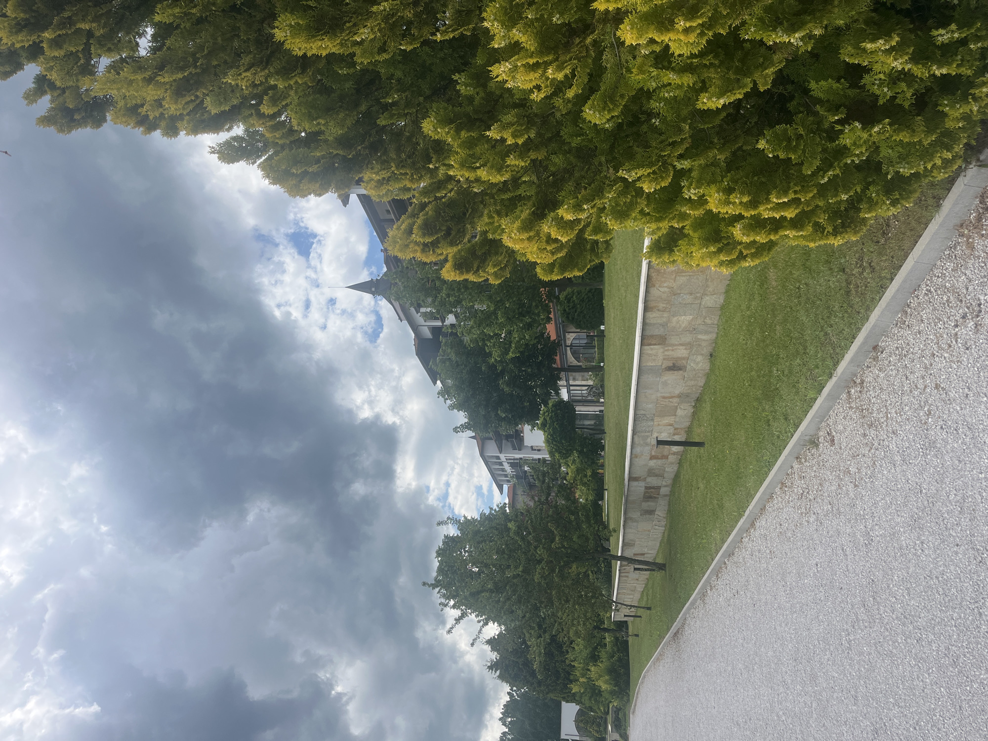 An outdoor scene in Razliv, Bulgaria, captured during the daytime under an overcast sky with heavy grey clouds and patches of brighter blue. The foreground features a wide, light-colored gravel path running along the right, bordered by a neatly mown green lawn. A stone retaining wall separates the lawn from a lower section on the left, with small, dark posts (possibly lights) visible within the grass.
In the middle ground, a variety of green trees and shrubs line the path and lawn. Behind these, several multi-story buildings with distinct traditional architecture are visible. These structures have dark, pointed roofs, light-colored facades, and numerous balconies, suggesting a resort or residential complex. A large, dense canopy of bright green trees, possibly conifers, frames the upper right portion of the image. No people are present, and there is no visible text. The scene conveys a serene atmosphere, typical of a well-maintained resort or park setting.