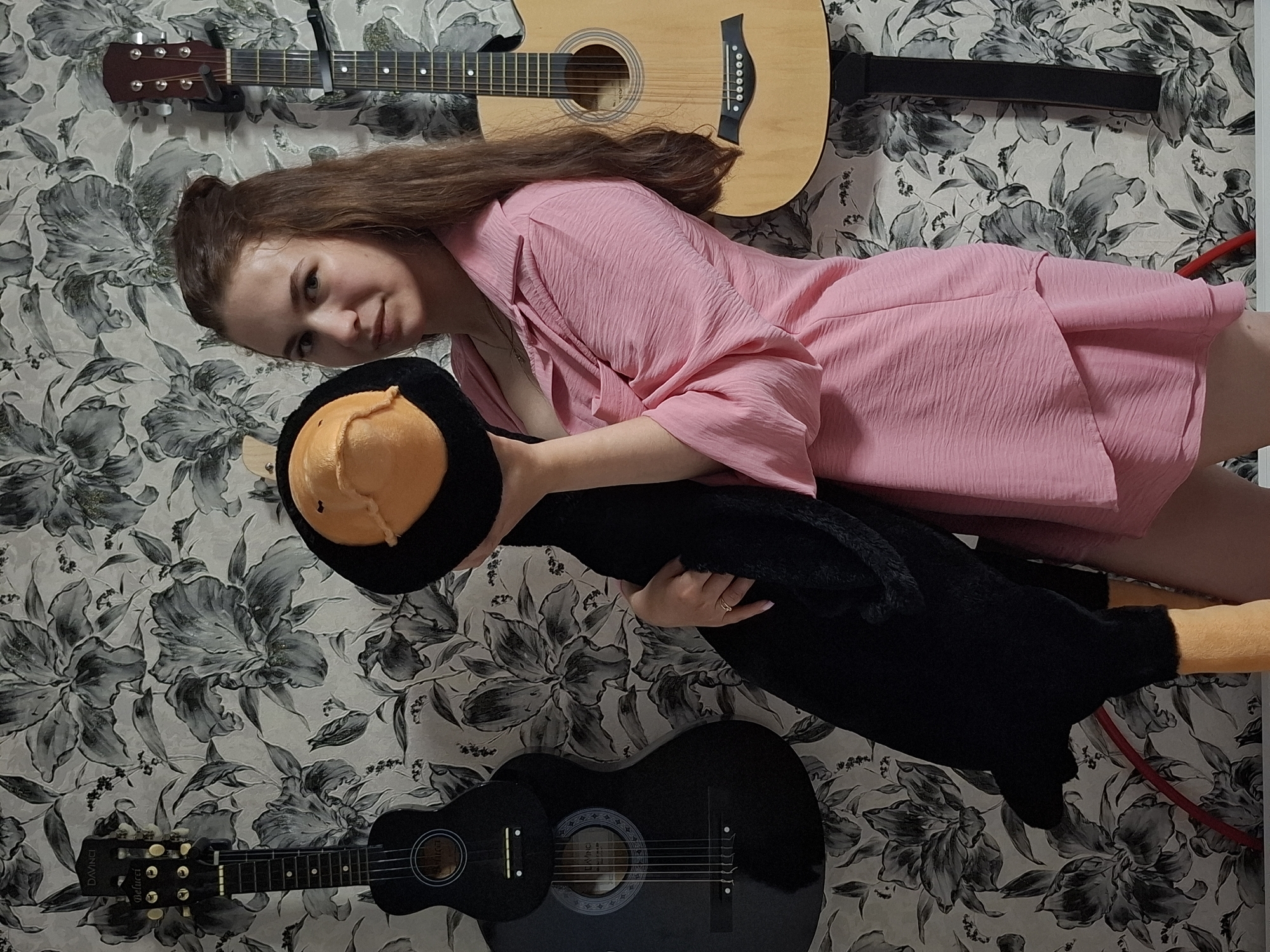 A woman is holding a penguin stuffed toy while lying down. Above her is an acoustic guitar, while below her is a black acoustic guitar. The background features a floral-patterned wall. This photo was taken at Chaikovsky, Perm Krai, Russia.