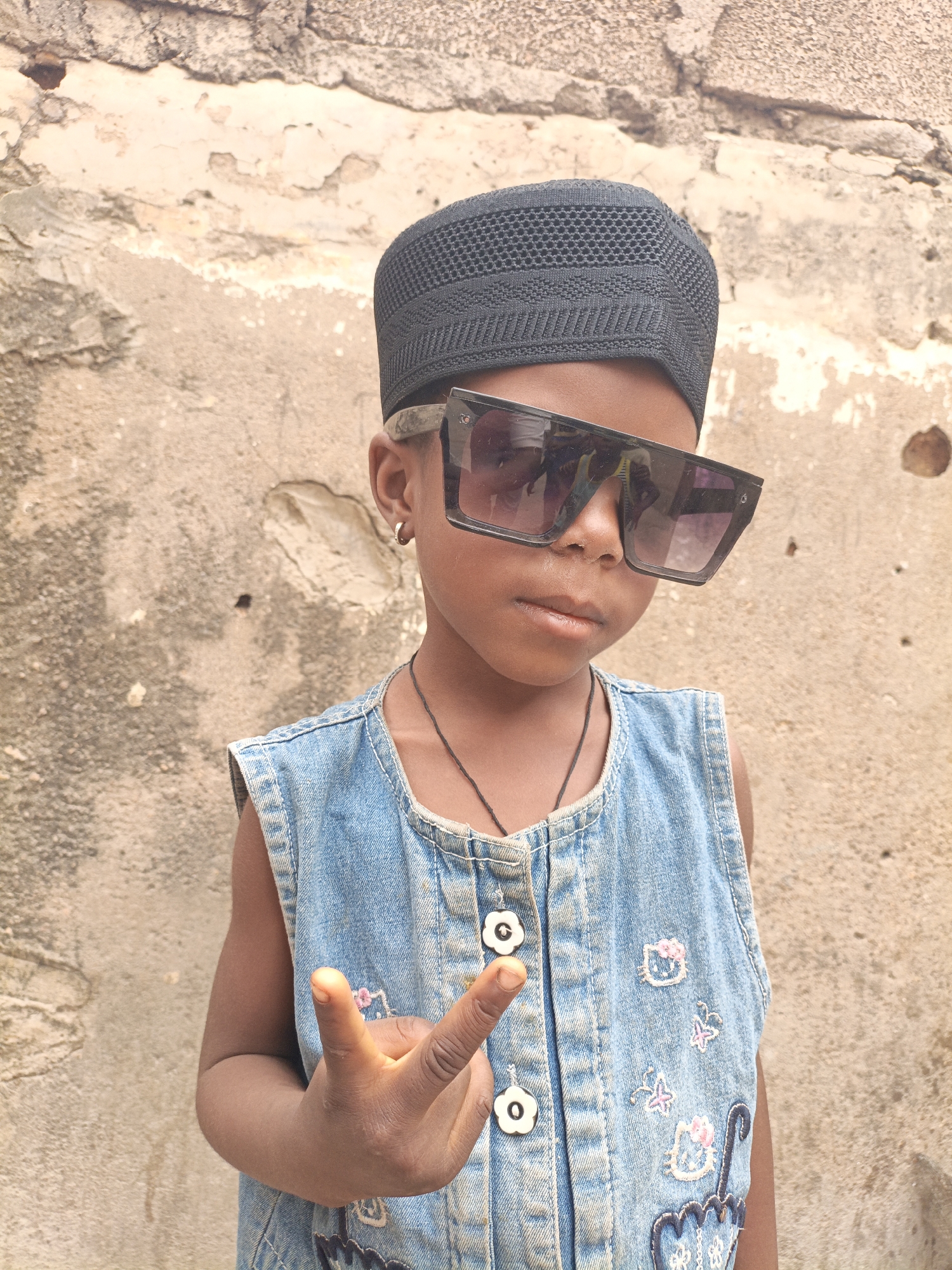 A young child stands in front of a textured beige wall. The child wears a black kufi cap, oversized sunglasses, and a sleeveless denim dress with decorative buttons and embroidered designs. The child is making a hand gesture, with the index and middle fingers extended. The location is Gwagwarwa, Nigeria, but there is no way to definitively identify any more details about the exact location based solely on the image provided.