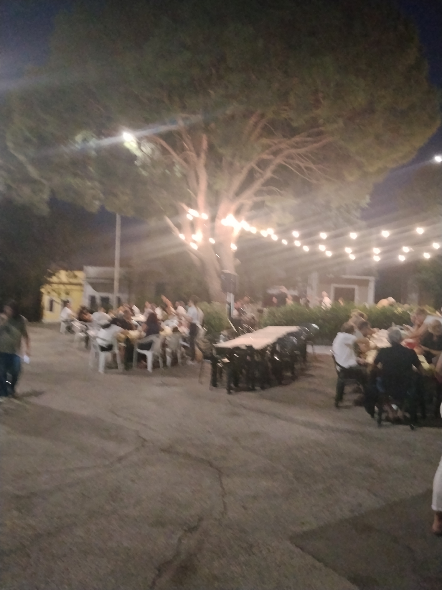 The image depicts an outdoor gathering at night, likely a celebration or event, under a large tree adorned with string lights. In the foreground, a paved area shows cracks and imperfections. To the left, a blurry figure of a person in a dark shirt walks away from the camera. In the midground, several round tables with white chairs are occupied by a group of people, some appearing to be eating or socializing. A longer, rectangular table with chairs is also visible, set for an event. In the background, there are low buildings with lights, and more people are milling about. The overall ambiance is festive and casual, suggesting a public event or a private party held outdoors. The city of Toritto, Italy, is mentioned as the location of this scene.
