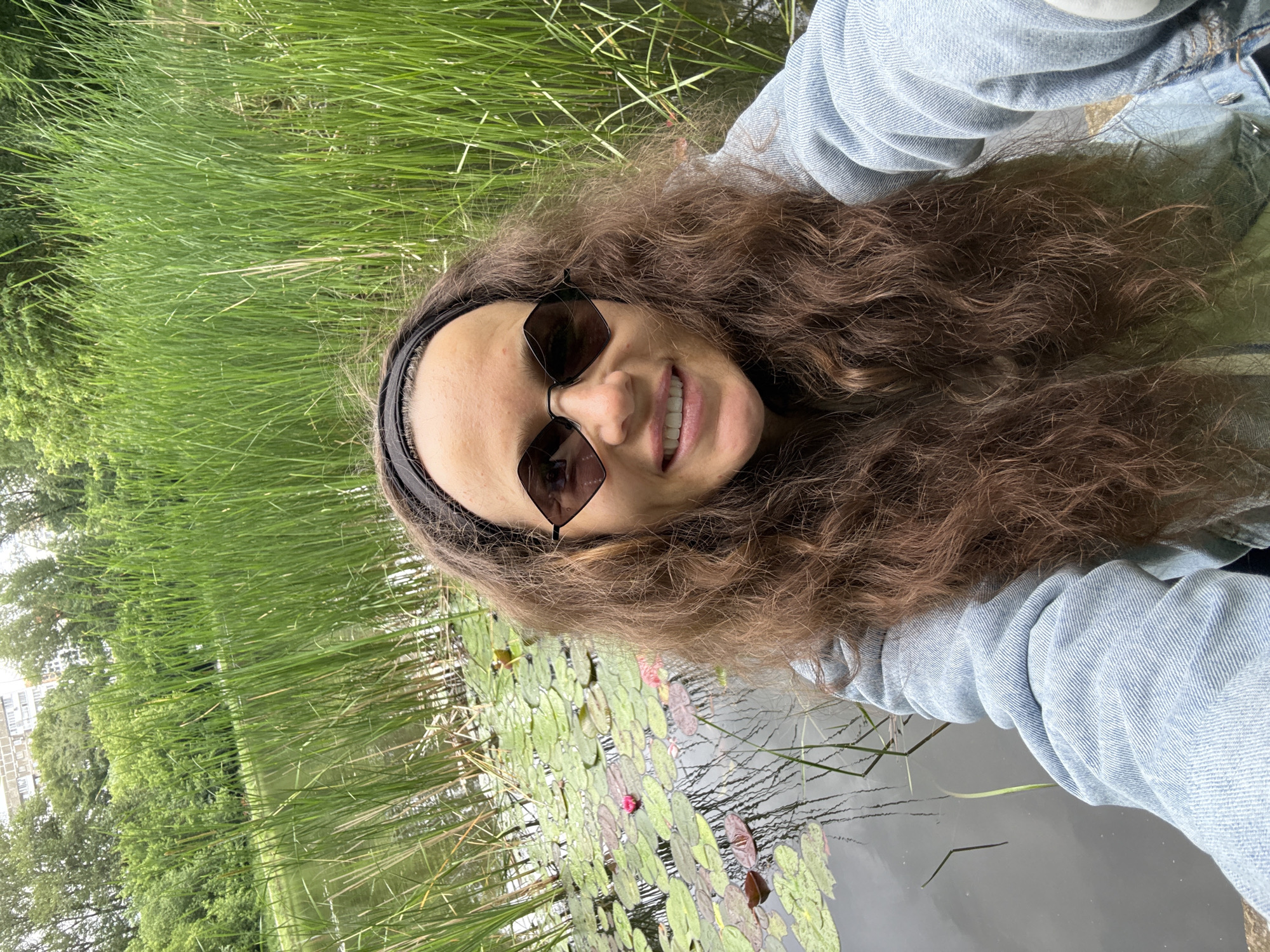 A woman is taking a selfie outdoors, in front of a body of water filled with lily pads and surrounded by tall grass. She is wearing a denim jacket, sunglasses, and a black headband. The background shows trees and a building. The location is a park in Moscow, Russia.