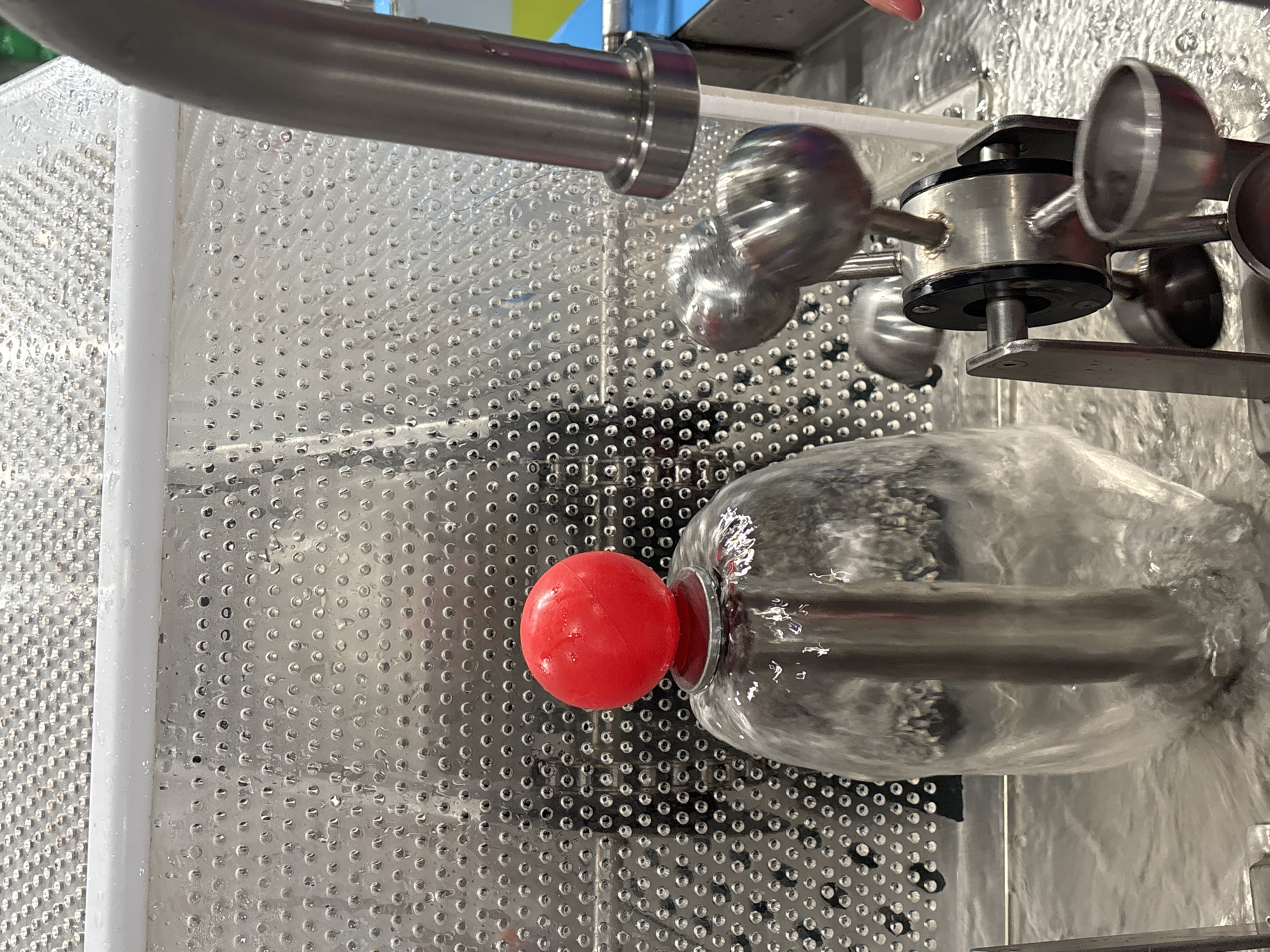 The image shows a close-up of a science exhibit or demonstration, possibly in Dubai, United Arab Emirates. The focal point is a clear, glass container filled with water, with a silver rod extending from the bottom and supporting a bright red ball at the top. Water is flowing around the rod and out of the container, creating ripples on the surface of the water in the surrounding area. To the right of the glass container is a complex metal apparatus with several spheres or balls, also appearing wet. In the background, a perforated metal sheet with a white bar across the top is visible, suggesting an indoor setting, possibly a museum or educational facility. The overall impression is a demonstration of fluid dynamics or a similar scientific principle.