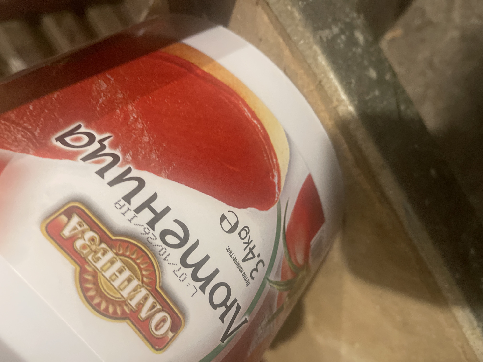 The image shows a large white plastic container, possibly a bucket, with a colorful label. The container is positioned at an angle, with its top surface facing the viewer. The label is predominantly red and white, featuring an illustration of what appears to be a red, circular food item, likely a tomato slice or a tomato-based product, and some green elements that might be leaves or stalks of tomatoes.
Visible text on the label includes:
- A brand logo in the bottom left, which reads "ВАЩЕЛИЕВО" (VASHCHELIEVO). The logo is set against a red background with a golden sunburst design.
- The product name, "Помидорица" (Pomidoritsa), which is written in large, stylized black Cyrillic letters with a white outline. This likely translates to "Tomato" or "Little Tomato".
- Below the product name, in smaller print, it says "НЕТТО КОЛИЧЕСТВО:" (Netto kolichestvo:), which means "Net quantity:".
- Adjacent to the net quantity, it specifies "3.4Kg e", indicating a net weight of 3.4 kilograms. The "e" symbol signifies conformity with EU weight and measure regulations.
- Further text, partially obscured and faded, appears to be a lot number or production date: "L:07/10/26/119".
The background of the image consists of blurry, dark, and textured surfaces, suggesting an indoor setting like a storage area or pantry, possibly with wooden or metal structures. No people are visible, and no activity is taking place. The lighting is somewhat dim, casting subtle reflections on the container's surface. The time of day is not discernible.