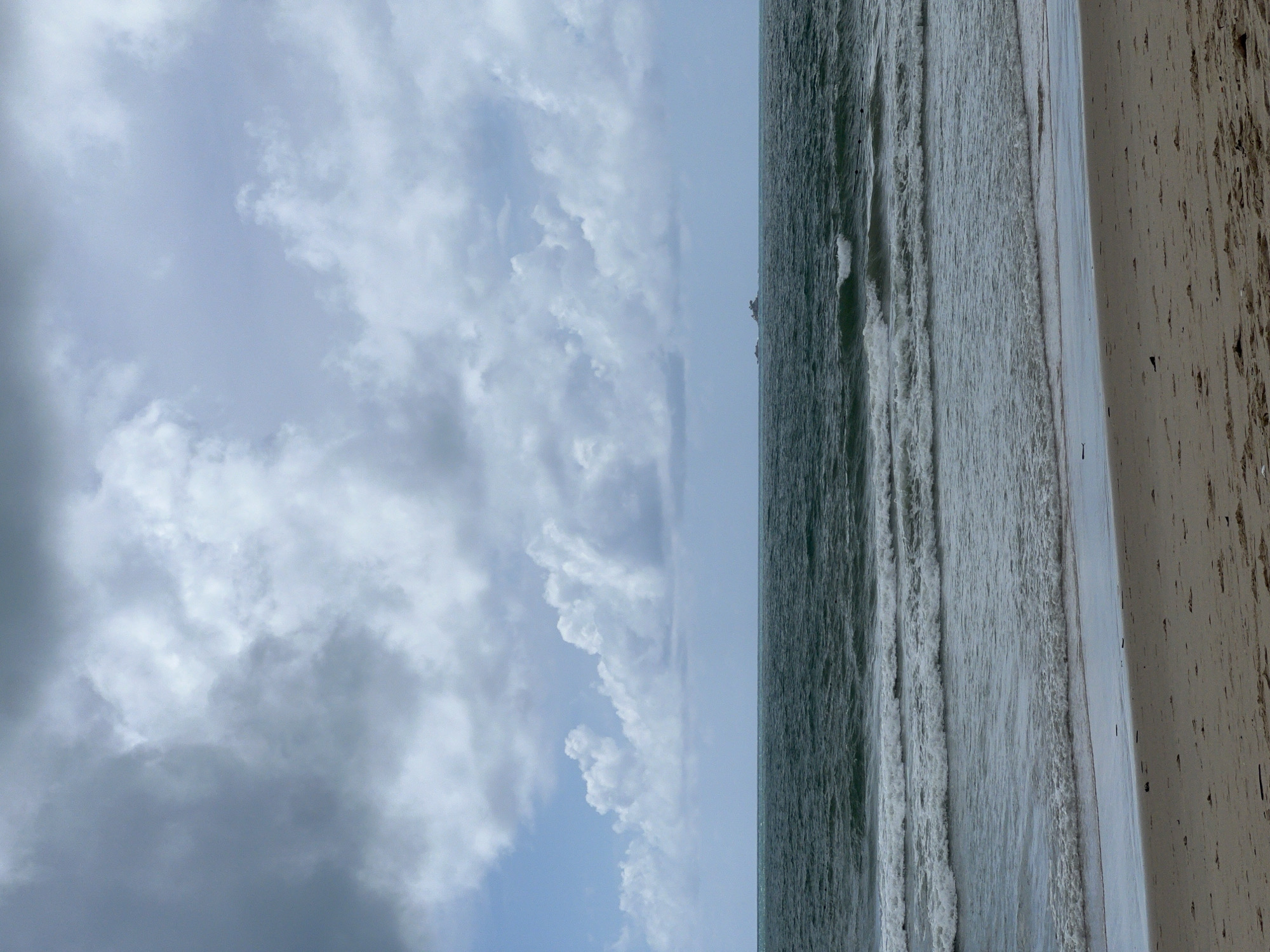 The image depicts a serene beach scene in Choeng Thale, Thailand, under a cloudy sky. The foreground features a sandy beach with scattered debris. The middle ground showcases the ocean with gentle waves breaking onto the shore. In the distance, two small islands are visible on the horizon, with a calm sea stretching out before them. The sky is filled with a mix of dark and light clouds, suggesting a possibly overcast or transitional weather condition. No people are visible in the frame.