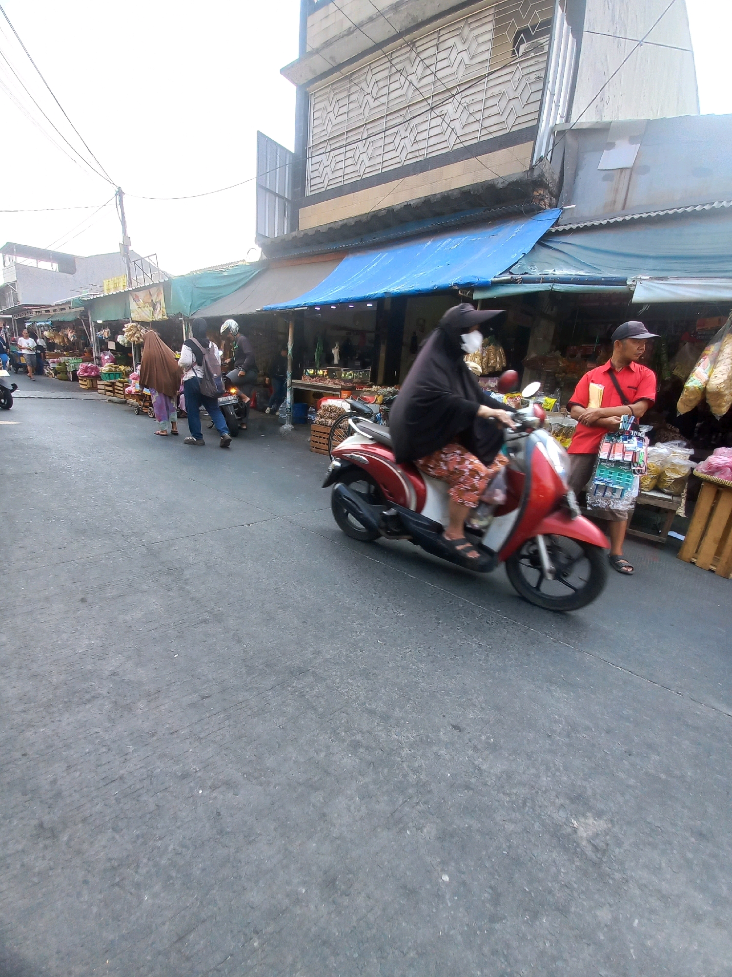 A low-angle, wide shot captures a street market scene in Depok, Indonesia. The foreground is dominated by a gray asphalt road, with a hint of a red and white scooter in the lower right corner. In the middle ground, a woman wearing a black hijab and a patterned skirt rides a red scooter. To her right, a man in a red shirt and black cap stands with a display of small items hanging from his arm. Behind them, market stalls line the street, covered by blue and green tarps. People are visible walking and browsing among the stalls. The background shows buildings and overhead wires. The overall atmosphere suggests a bustling market day.