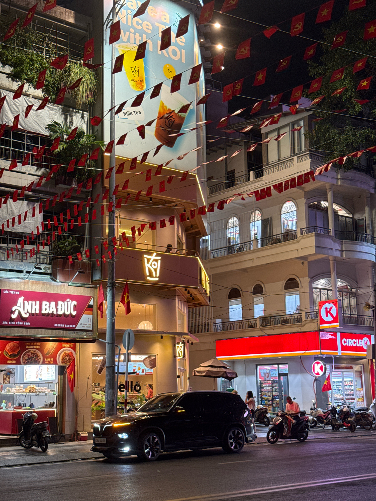 The image depicts a street scene at night in Nha Trang, Vietnam, featuring a row of buildings decorated with numerous small red flags. The flags, bearing a yellow star and hammer and sickle emblem, suggest a patriotic or political display. On the left, a building has a prominent red sign that reads "Anh Baduc" with smaller text below, indicating a business. Adjacent to it, a store with a bright yellow logo that appears to be "KOI The" is visible, advertising milk tea. Further to the right, a Circle K convenience store is illuminated with its distinctive red and white branding. A black car is parked in the foreground on the right side of the street, and several motorcycles are parked along the sidewalk in front of the businesses. People are visible walking and standing near the stores. The buildings are multi-story, with balconies and arched windows, typical of urban architecture. The overall atmosphere is illuminated by streetlights and the glow from shop signs.
