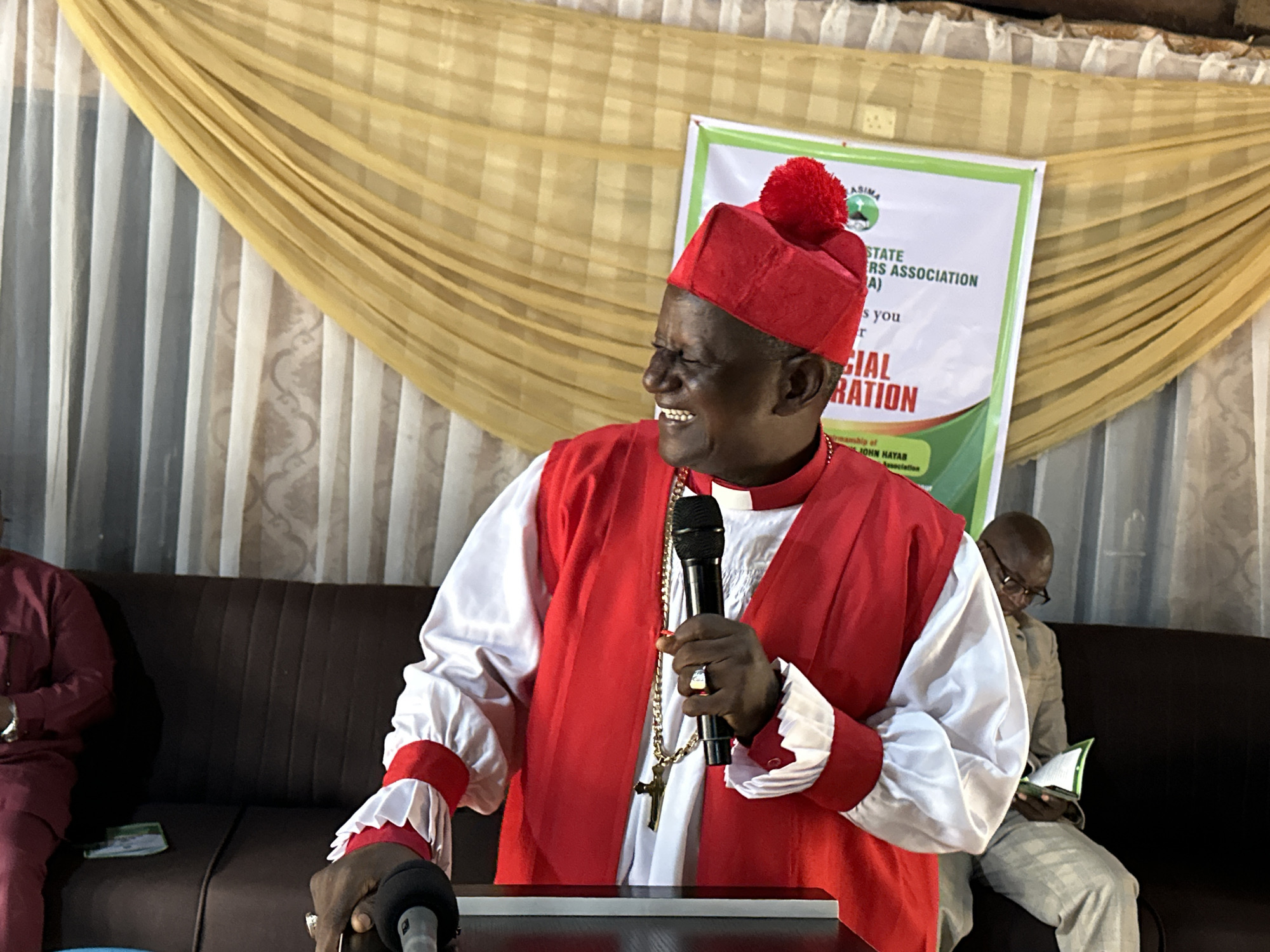 In a room in Barden Yana, Nigeria, a Black man wearing a red and white religious garment and a red hat with a pom-pom is speaking into a microphone. He is holding the microphone in his right hand and has a gold chain with a cross around his neck. The man is smiling and his eyes are closed. Behind him, a banner reads "ASIMA STATE TRS ASSOCIATION". To the left of the main subject, another man in a maroon outfit is sitting on a brown couch. To the right of the main subject, a man wearing glasses and a tan suit is sitting on the same couch and reading a book. The room is decorated with yellow and white curtains.