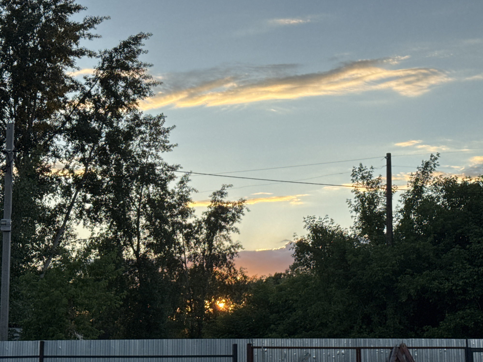 The image captures a serene sunset scene in Chaikovsky, Russia, characterized by a soft, warm sky and silhouetted trees. The dominant feature is the sky, transitioning from a light blue-grey at the top to warmer hues of pale yellow, orange, and soft pink towards the horizon, particularly on the right side. Elongated, thin clouds, illuminated by the setting sun, stretch across the sky, displaying a golden glow.
In the midground and foreground, lush, dark green trees stand out as prominent silhouettes against the brighter sky. The sun is partially visible, peeking through the dense foliage towards the lower-center right, casting a bright, golden-orange light that backlights some of the leaves. Two utility poles are visible: one on the far left, partially obscured by trees, and another on the right side, centrally positioned among the trees. Thin electrical wires stretch horizontally across the frame, connecting these poles and passing through the tree canopy.
The very bottom of the frame is occupied by a corrugated metal fence, appearing in shades of grey and black, suggesting a boundary or property line. The overall impression is one of a peaceful evening, with the natural beauty of the sunset contrasting with the man-made structures of the fence and utility lines. There is no visible text in the image.