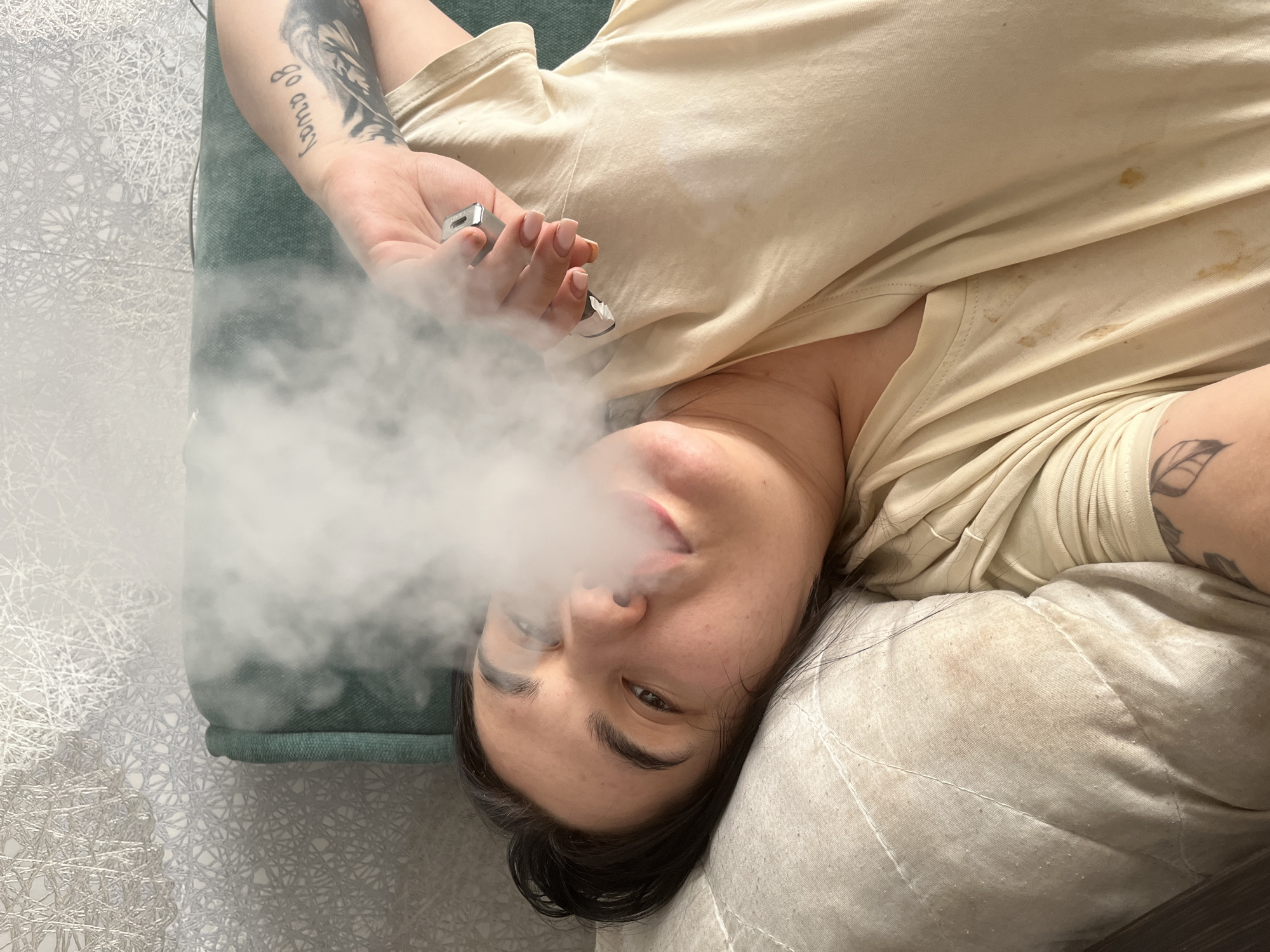 A person is lying on a pillow with smoke billowing from their mouth, holding a vape device. They are wearing a light-colored T-shirt. Tattoos are visible on their arms. The setting appears to be indoors, with a decorative surface visible in the background. There is no information to definitively identify the location as Chaikovsky, Russia.