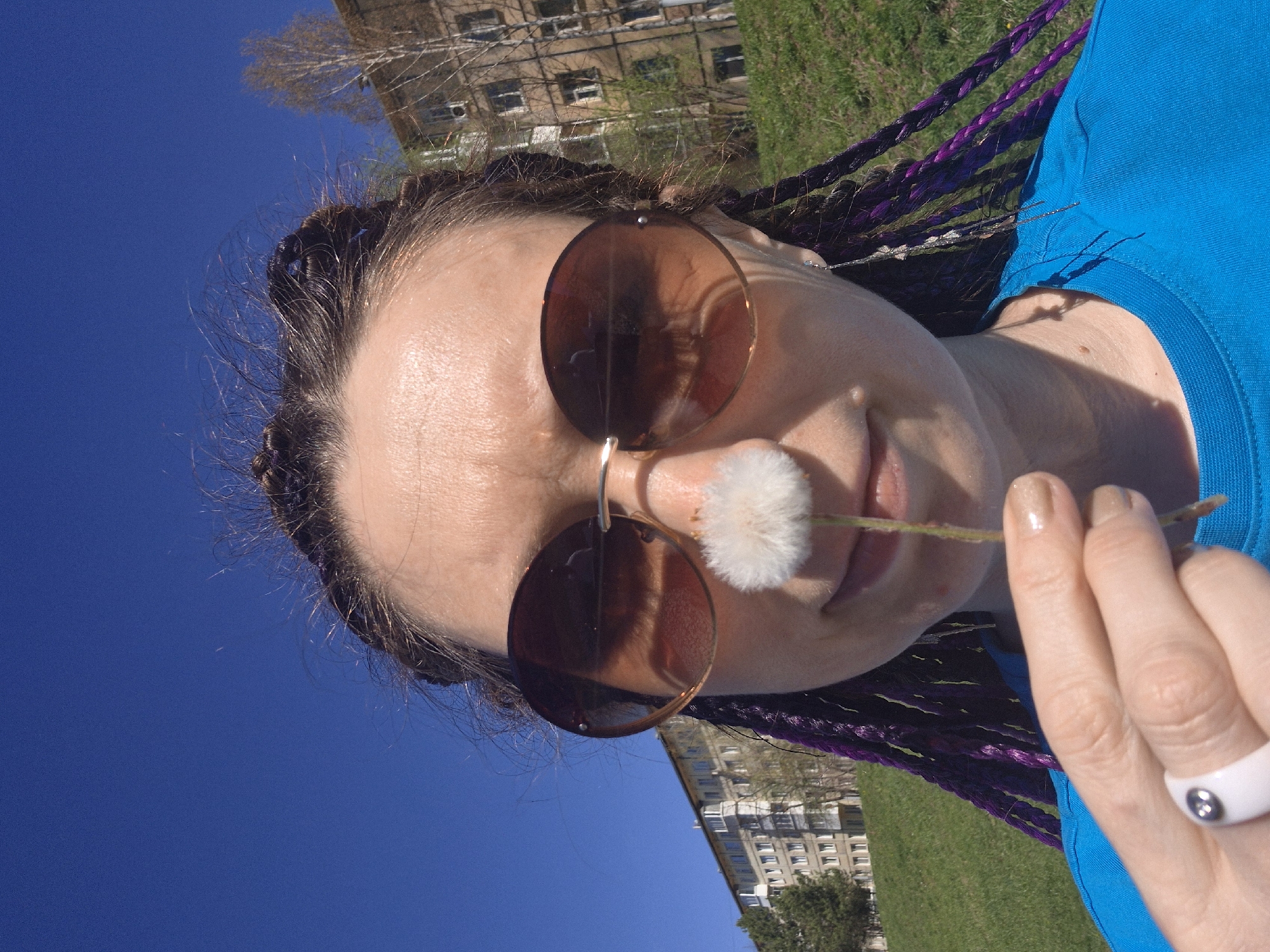 A young woman wearing sunglasses and a blue shirt is holding a dandelion in front of her mouth, with her hand visible and adorned with a white ring. Her hair is styled in braids with purple strands. The background features an apartment building and grassy area, with a bright blue sky overhead. The image appears to have been taken outdoors in an urban setting resembling Votkinsk, Russia, considering the typical architecture of the apartment buildings in the background.