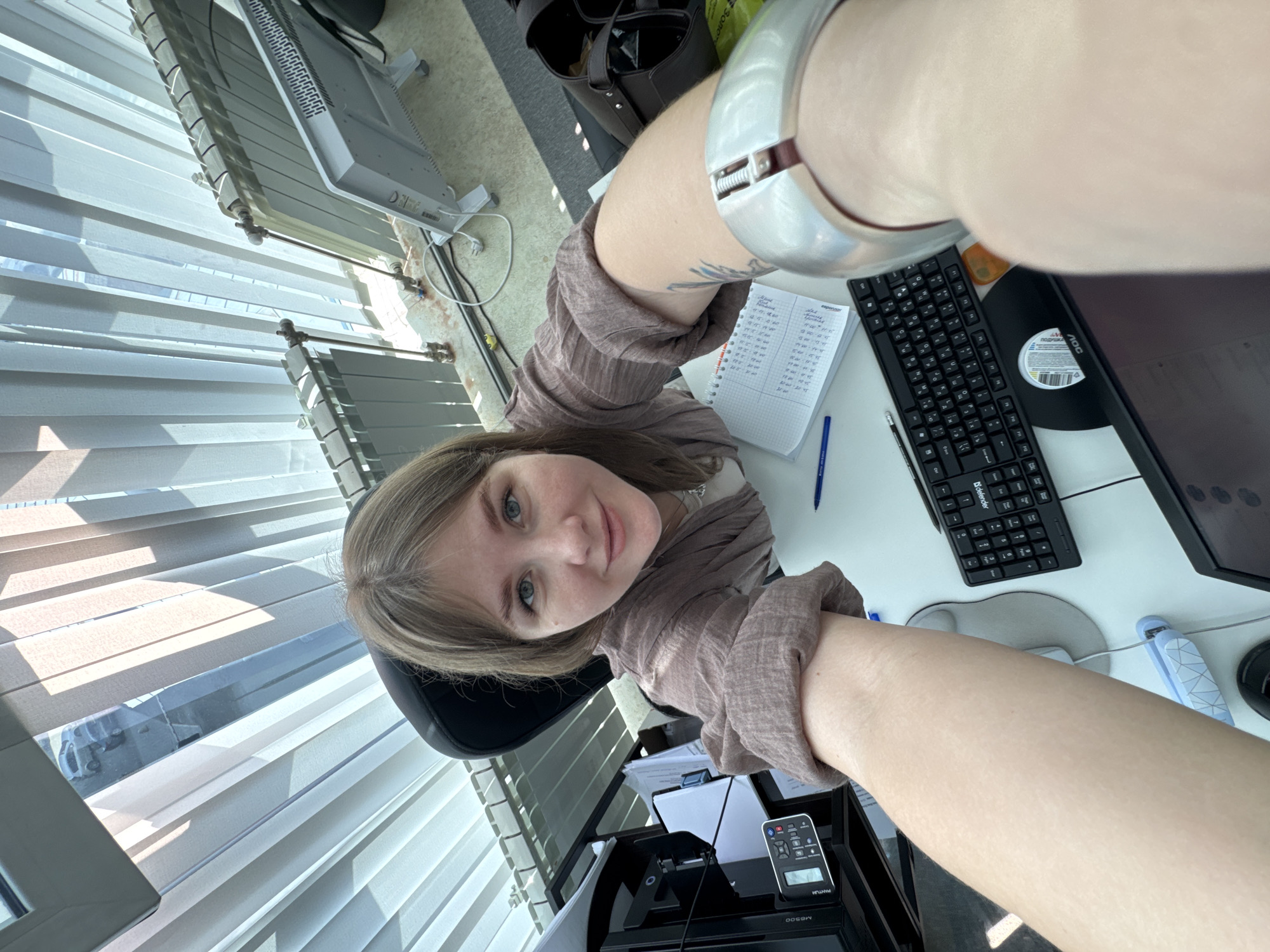 A woman with light brown hair and fair skin is taking a selfie in an office. The office has blinds on the window, a computer monitor, keyboard, mouse, stapler, notebook with handwritten text, pen, and a printer/scanner. The office is likely located in Perm, Russia, as indicated by the text "ПОДГОТОВКА" (preparation) written on the round sticker on the computer monitor in Russian. The woman is wearing a brown shirt and a silver bracelet.
