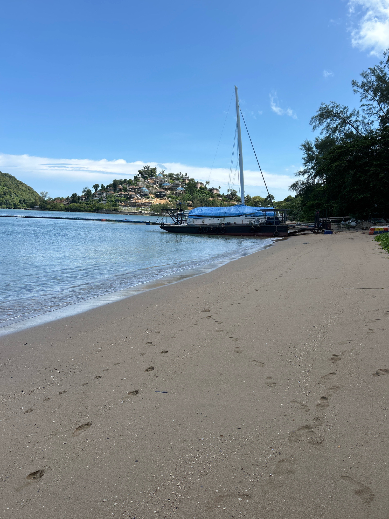 The image depicts a scenic view of a beach in Ratsada, Thailand. The foreground is dominated by a sandy beach with visible footprints leading towards the water. The sea is calm and reflects the clear blue sky, with gentle waves lapping at the shore. In the mid-ground, a stationary boat with a blue tarp covering its deck is moored near the beach. The boat has a tall mast, suggesting it's a sailboat. Further in the background, a lush green hillside is dotted with houses and buildings, creating a picturesque view of the coastal settlement. Trees line the edge of the beach and the hillside, adding to the natural beauty of the scene. The sky is bright blue with a few scattered clouds.