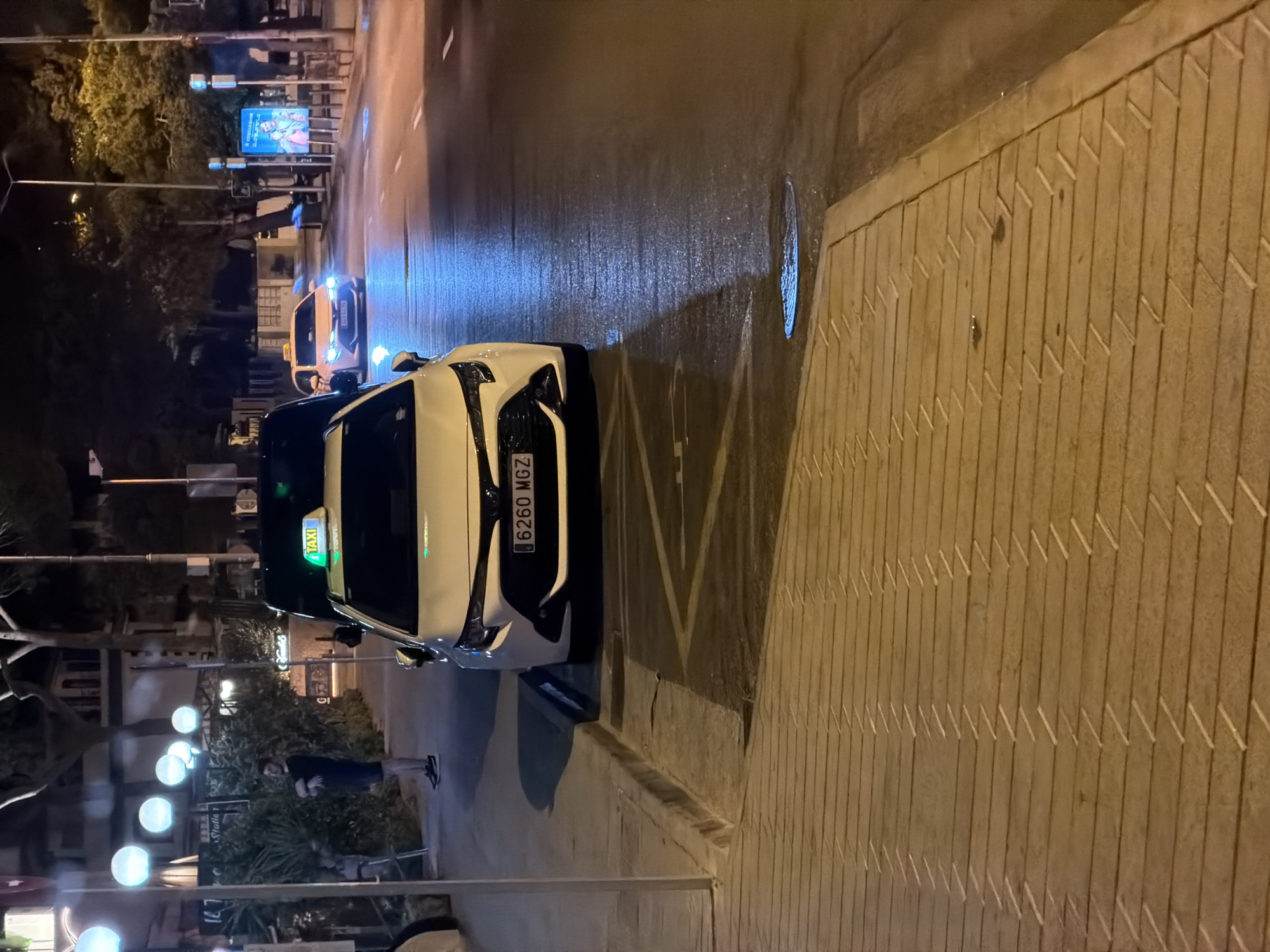 At night, a white Toyota Corolla taxi with license plate "6260 MGZ" is parked on the side of a street in Palmanova, Spain. The taxi has a lit "TAXI" sign on its roof. The street is wet and reflects streetlights and the headlights of another taxi, a yellow Toyota, driving in the distance. A man in a dark t-shirt and shorts is standing on the sidewalk to the left of the taxi, with his arms crossed. The sidewalk is paved with light brown rectangular tiles arranged in a herringbone pattern. In the background, there are trees, buildings with lights, and decorative spherical lights hanging from poles.