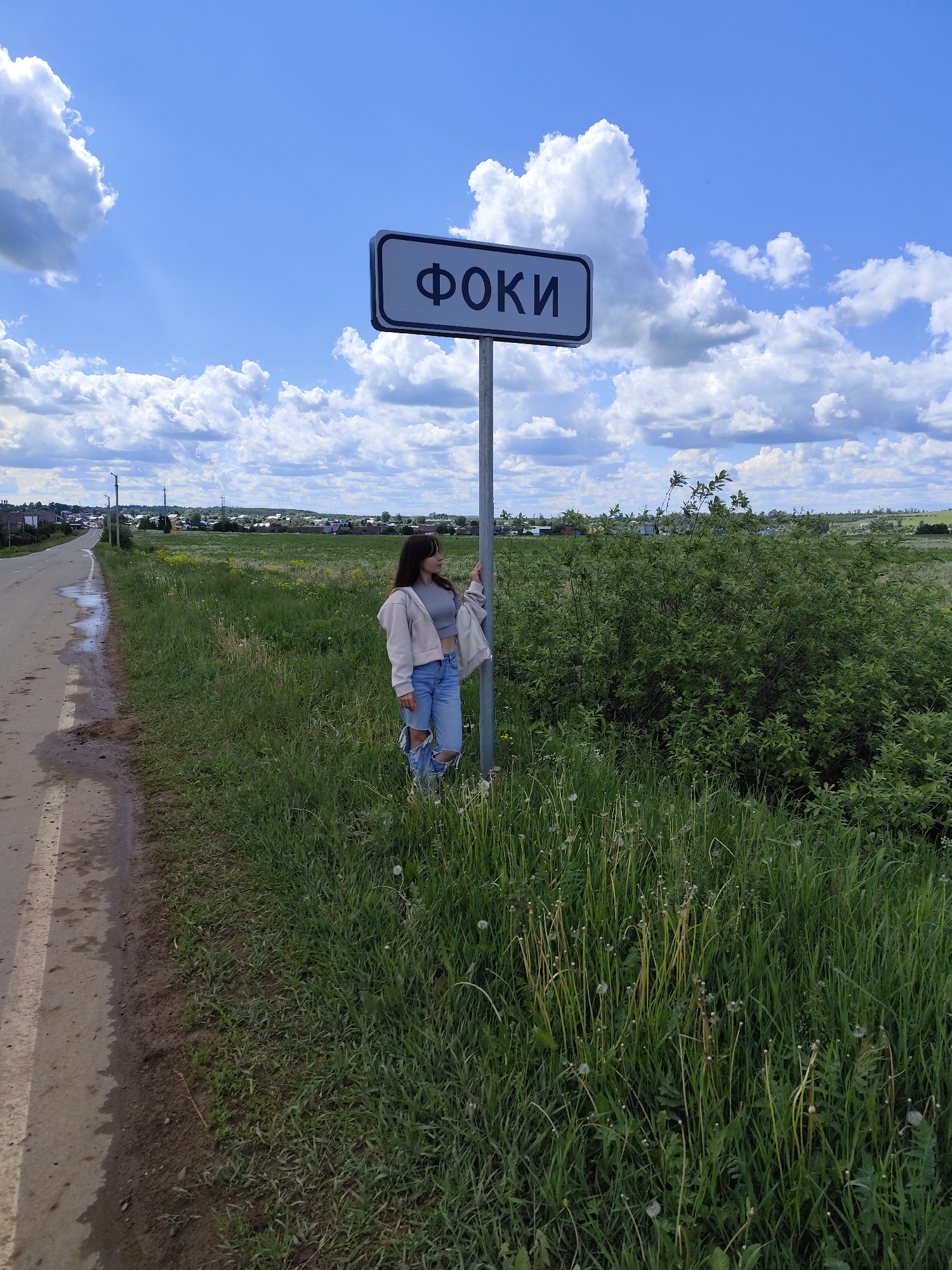 A full shot depicts a young woman standing by a sign post in Фоки, Russia. She is dressed in a gray top, ripped blue jeans, and a light-colored jacket. She leans against the sign post, which displays the name of the city in Cyrillic letters.
The scene is set in a rural area next to a paved road with visible water puddles. The background features a field of green grass and vegetation, with a line of trees and houses in the distance. The sky is blue with scattered white clouds, indicating a sunny day. The sign post is positioned in a patch of tall grass and dandelions.