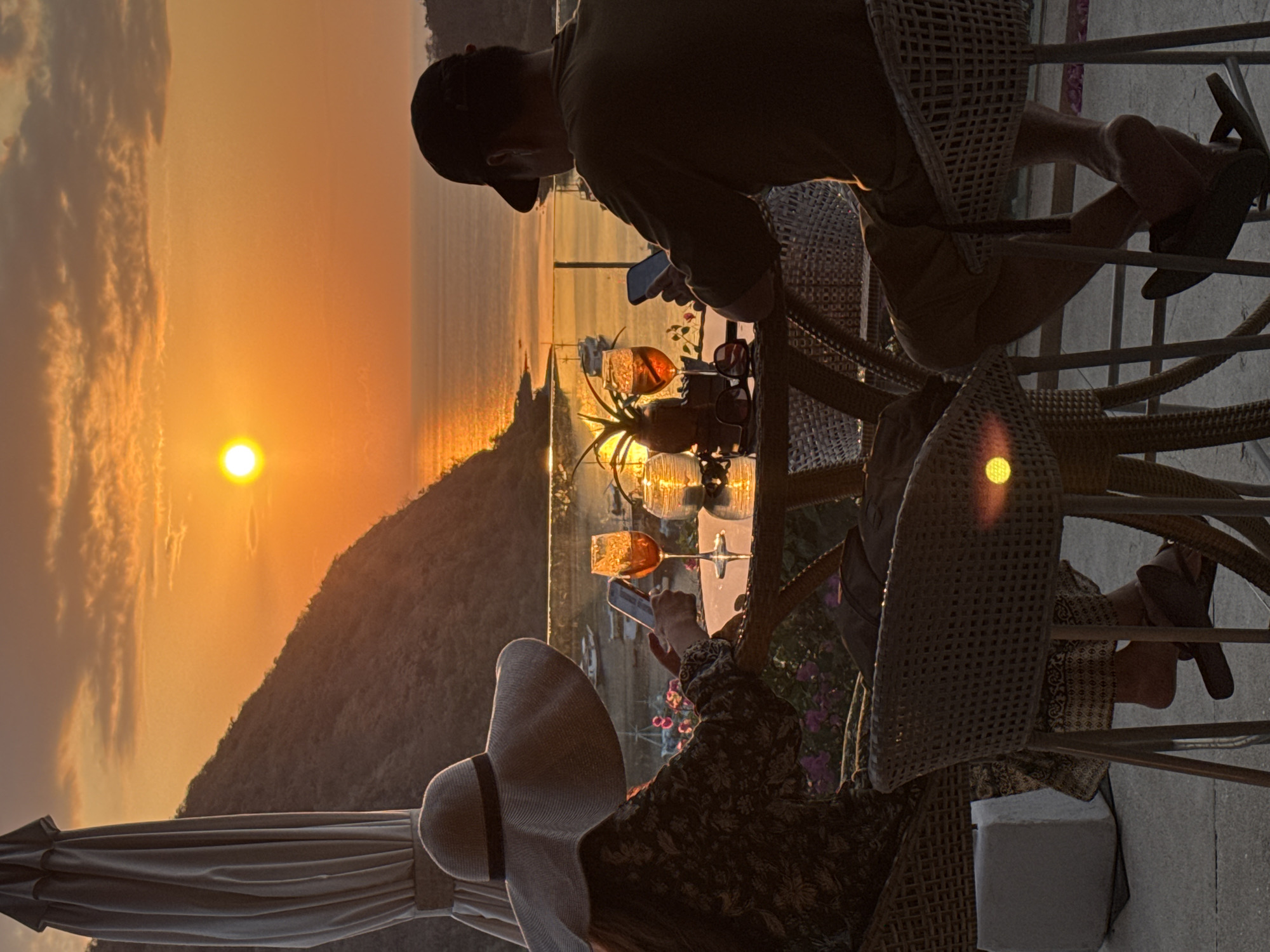 At sunset in Labuan Bajo, Indonesia, two individuals sit at a table on an outdoor patio. The sky is a vibrant orange, with the sun low on the horizon casting a golden glow over the ocean and surrounding hills. A large umbrella is positioned to the left of the frame, casting a shadow over a woman wearing a wide-brimmed hat and a patterned dress. She is looking down at her phone, which she holds in her right hand. In front of her is a table with two glasses of an amber-colored beverage, a small potted plant, and a lit candle. To her right, a man wearing a baseball cap and a dark t-shirt also looks at his phone. He is seated in a woven chair, with his legs crossed. The table is made of rattan material, and the chairs are also woven. In the background, the ocean stretches out, with a few boats anchored in the distance. The scene suggests a relaxed evening, with the individuals enjoying the sunset and their drinks.