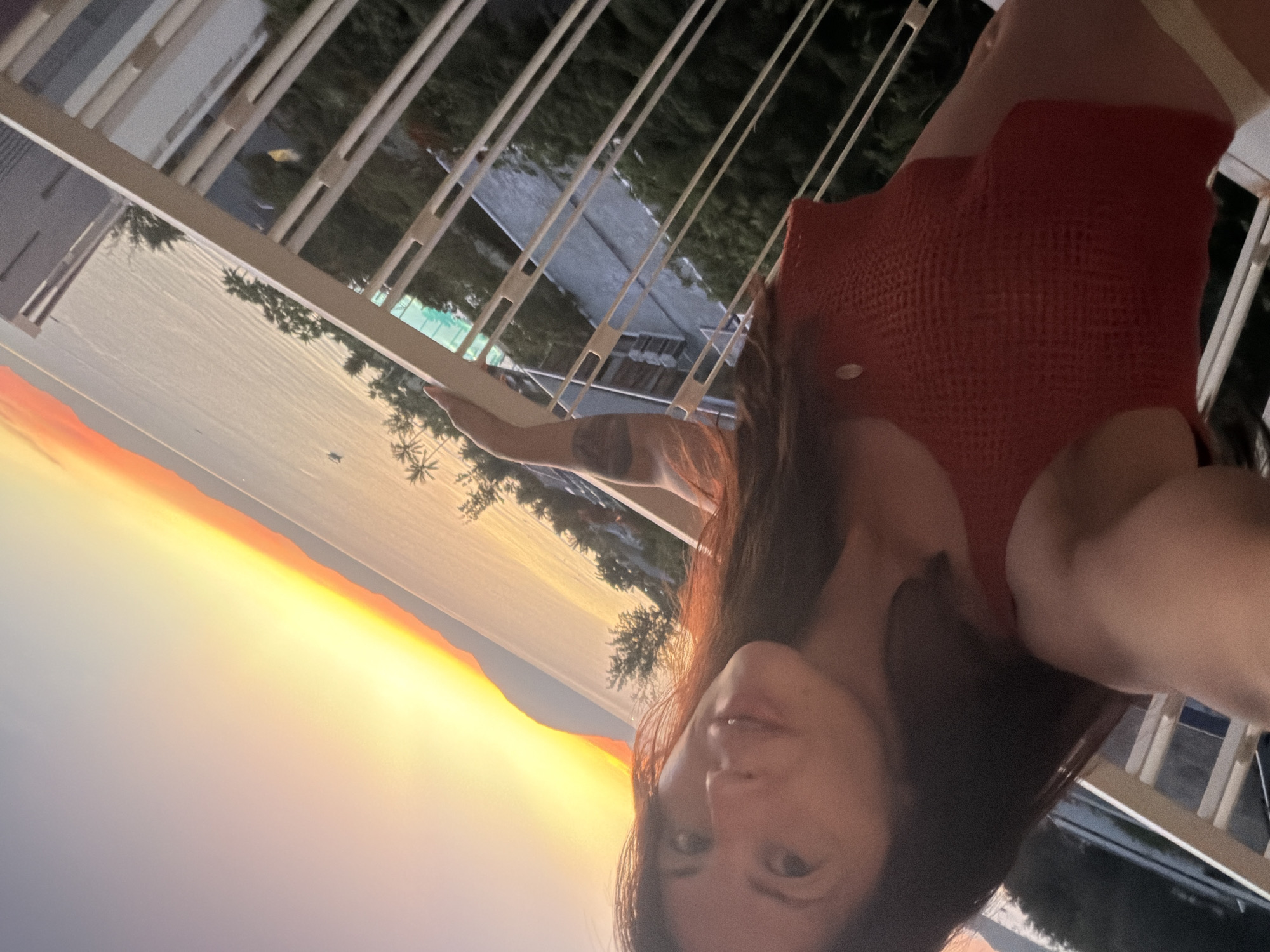 In this portrait-style, over-the-shoulder shot, a woman with long, dark hair and a red knit top is captured from a high vantage point on a balcony, looking out at a sunset over the ocean. The sky transitions from a soft blue to a vibrant orange and yellow gradient. In the distance, a coastline with mountains is visible, and the calm sea is dotted with a few small boats. Palm trees line the shore. The balcony railing, made of white metal bars, runs diagonally across the frame, with the woman's arm resting on it. Buildings and lush green foliage are visible in the background, suggesting an urban or resort setting. The overall atmosphere is serene, with the golden hues of the sunset creating a warm and inviting ambiance. It is assumed this location is Ninh Hòa Ward, Đồng Nai Province, Vietnam due to the geographical context of Ninh Hòa Ward being a coastal area in Vietnam known for its beaches and natural beauty.